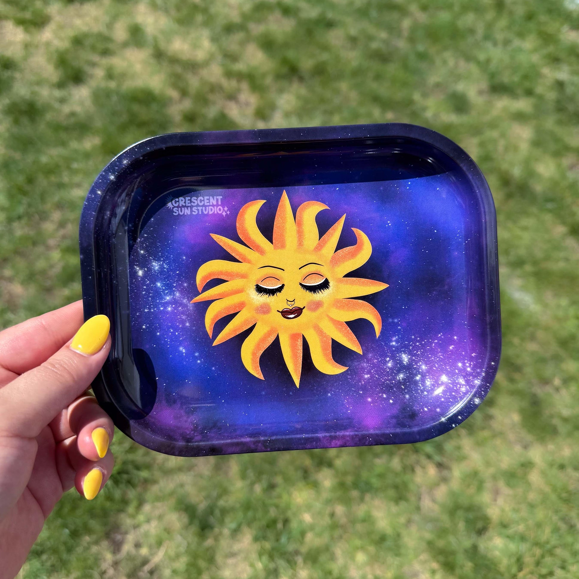 This rectangle tray with raised edges, features a blissed out and elevated glam sun floats in a purple-blue galaxy background. This personified sun has thin eyebrows, closed eyes with full lashes and eyeshadow, blushing cheeks, and glossy full lips.

A hand with yellow nails is holding the tray up to the camera against a green textured background.