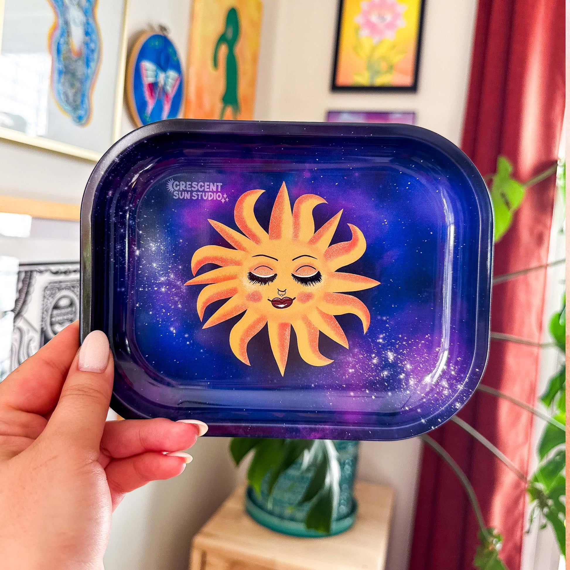 This rectangle tray with raised edges, features a blissed out and elevated glam sun floats in a purple-blue galaxy background. This personified sun has thin eyebrows, closed eyes with full lashes and eyeshadow, blushing cheeks, and glossy full lips.

This image shows the scale of the tray, as it is held up to the camera in a person's hand. In the background of the photo is an art gallery wall.