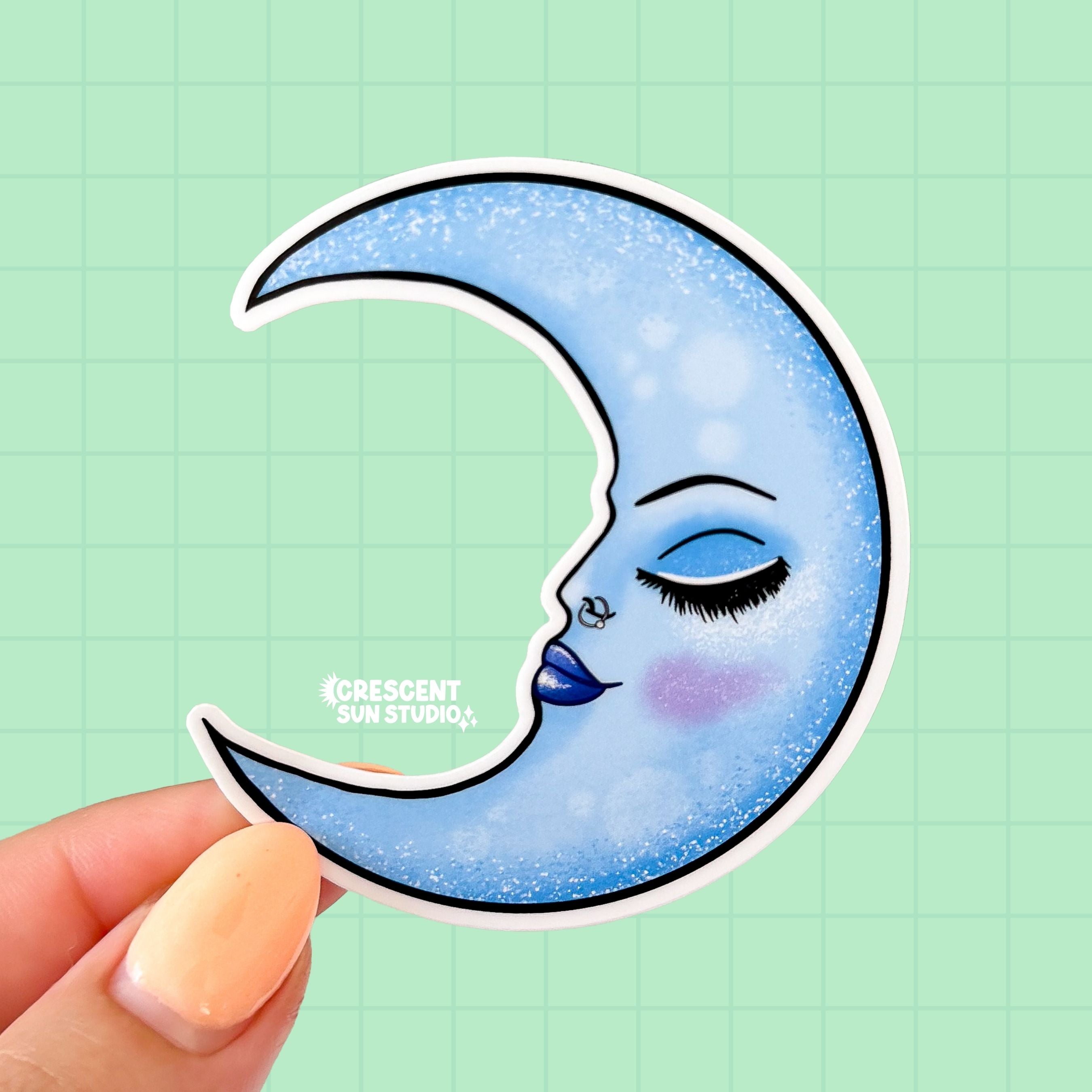A sticker in the shape of a crescent moon. The sticker is shaded blue and has the profile of a serene and confident woman's face. The face has a thin arched eyebrow, closed eye with deeper blue shadow and lush lashes. She has flushed cheeks and full lips that are glossy purply-blue.