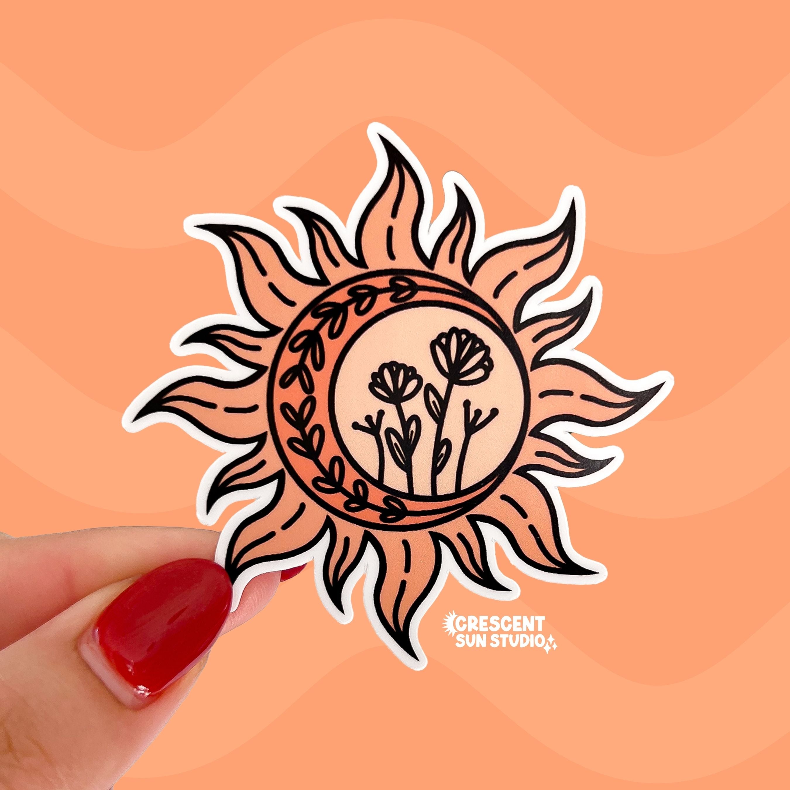 A pale orange die cut sticker in the shape of the sun with dancing rays. Inside the circle of the sun is a darker orange crescent and black doodles of wildflowers and vines.