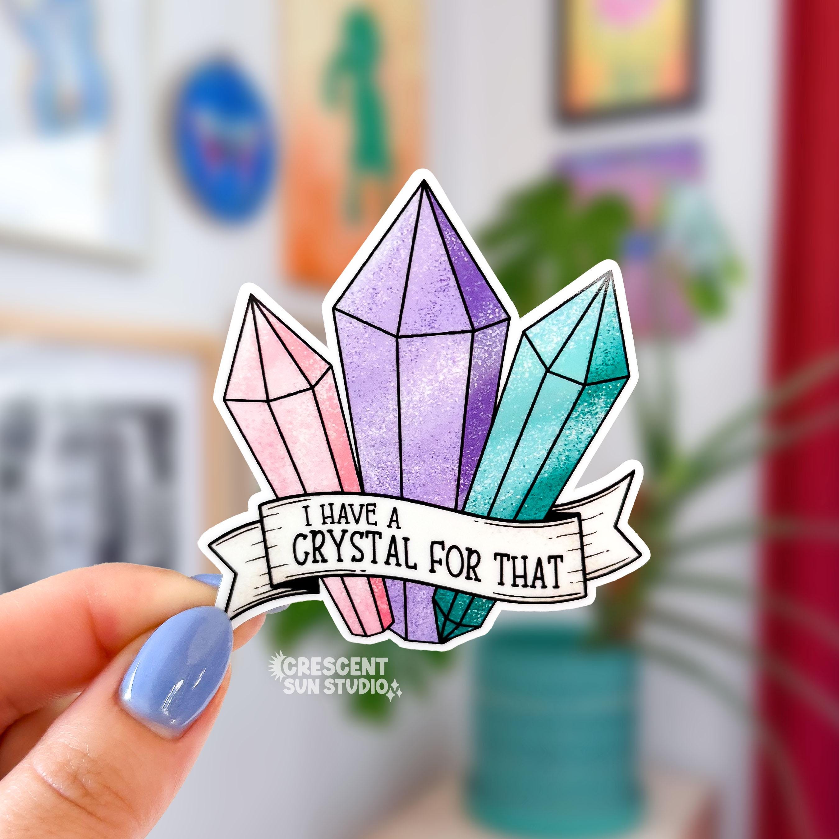 A sticker in the shape of three pastel shaded crystals in pink, purple, and turquoise. At the bottom is a white banner with black text that reads "I have a crystal for that"