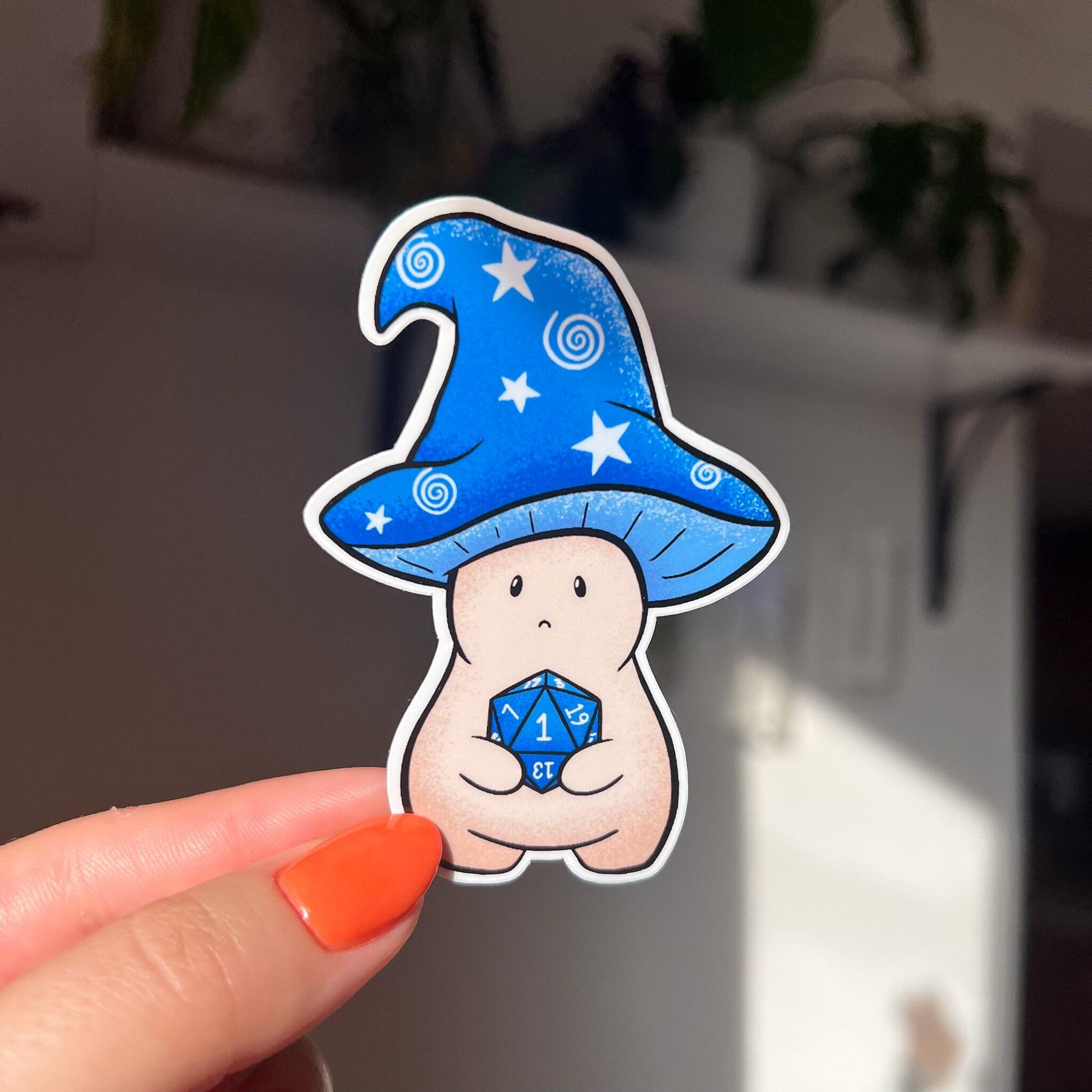 A sticker in the shape of a wizard mushroom character. The mushroom cap is blue with white stars and spirals. The magician holds a 20-sided die, used in tabletop games, and has rolled a 1. You can tell from his face that he's sad about it.