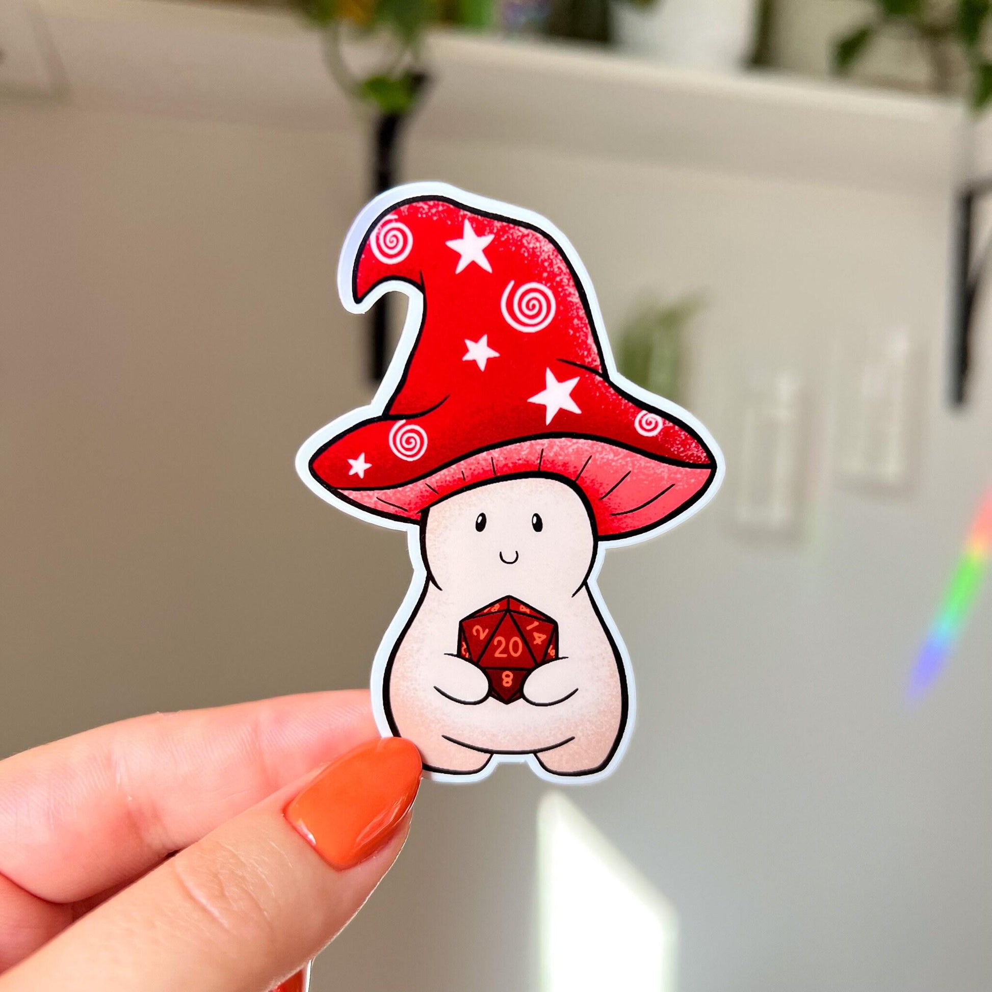 This sticker is shaped like a happy mushroom character whose cap is shaped like a red wizard's hat holds a d20 die. In this image, a hand holds the sticker close to the camera.