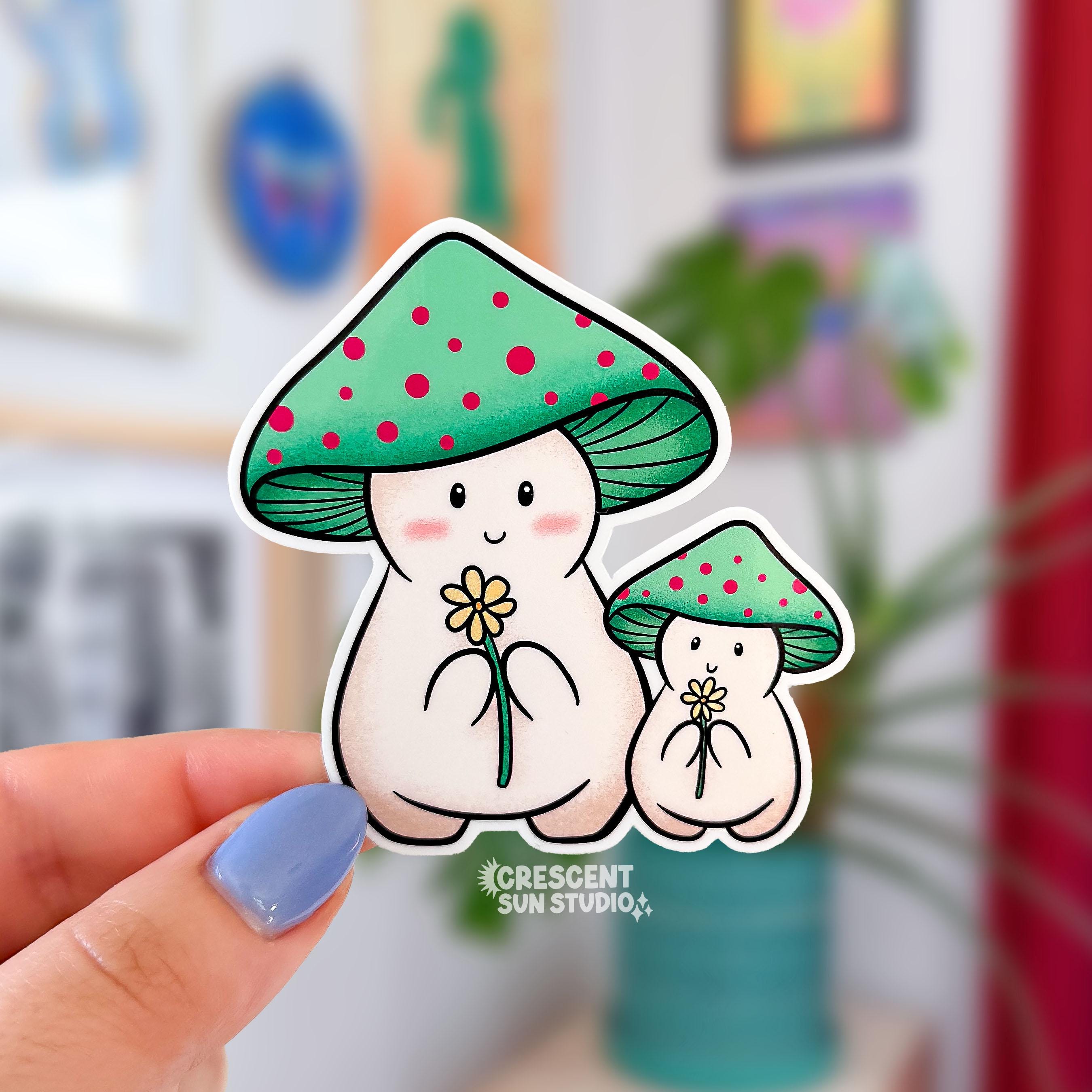 A sweet die cut sticker of two mushroom characters. Both have green mushroom caps with red spots. They are both light beige and holding a yellow daisy. One mushroom is smaller than the other, indicating a child. 