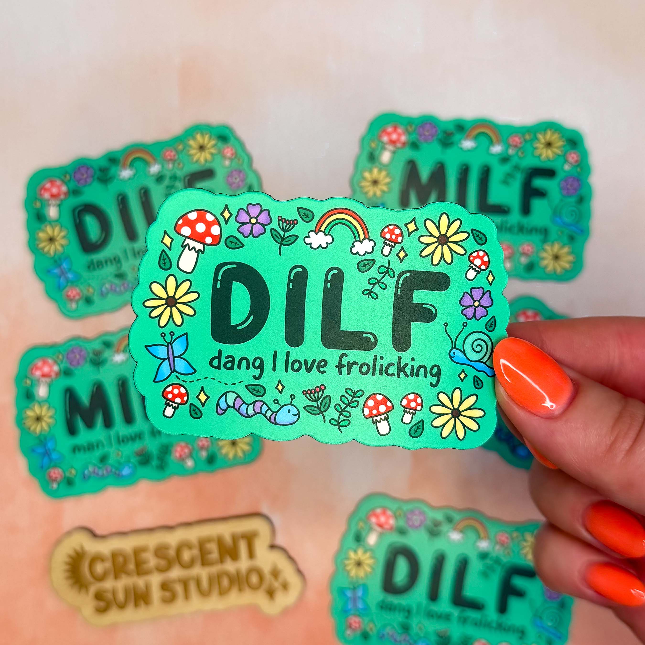A bright green colored rectangle magnet with black bubble letters that says DILF and in smaller letters underneath that reads "dang, I love frolicking." The text is bordered by brightly colored mushrooms, rainbows, butterflies, and various flowers.

In this image, the magnet is held close to the camera with other magnets slightly blurred in the background.