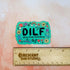 A bright green colored rectangle magnet with black bubble letters that says DILF and in smaller letters underneath that reads "Dang, I love frolicking." The text is bordered by brightly colored mushrooms, rainbows, butterflies, and various flowers.

In this image, the magnet sits above a wooden ruler, showing its size of 3 inches.