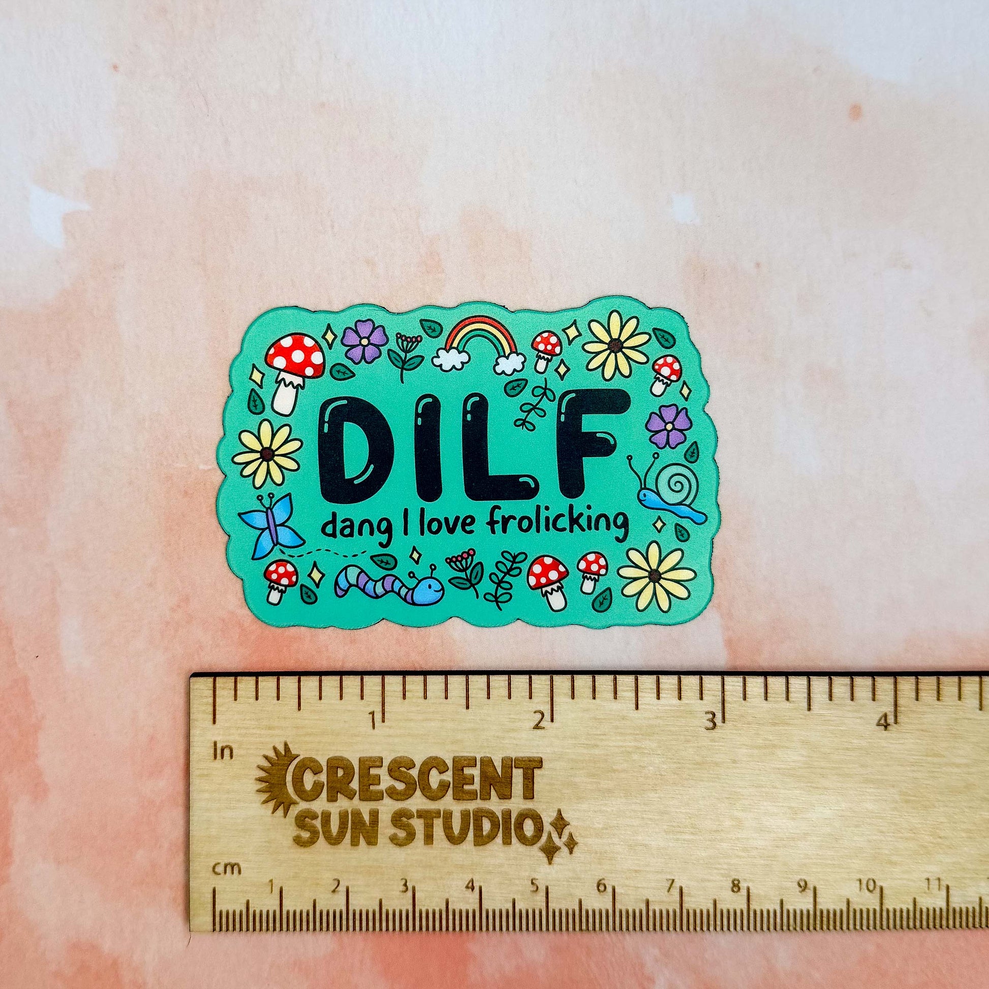 A bright green colored rectangle magnet with black bubble letters that says DILF and in smaller letters underneath that reads "Dang, I love frolicking." The text is bordered by brightly colored mushrooms, rainbows, butterflies, and various flowers.

In this image, the magnet sits above a wooden ruler, showing its size of 3 inches.