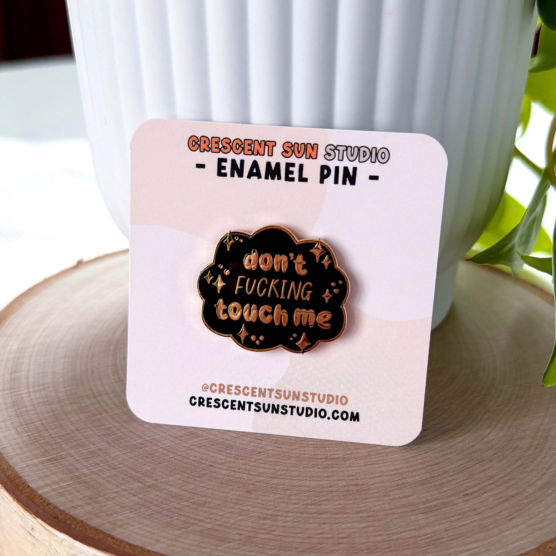 A black cloud shaped pin with gold lettering that reads "don't fucking touch me" surrounded by little stars. In this image, the pin is displayed on a light colored card.