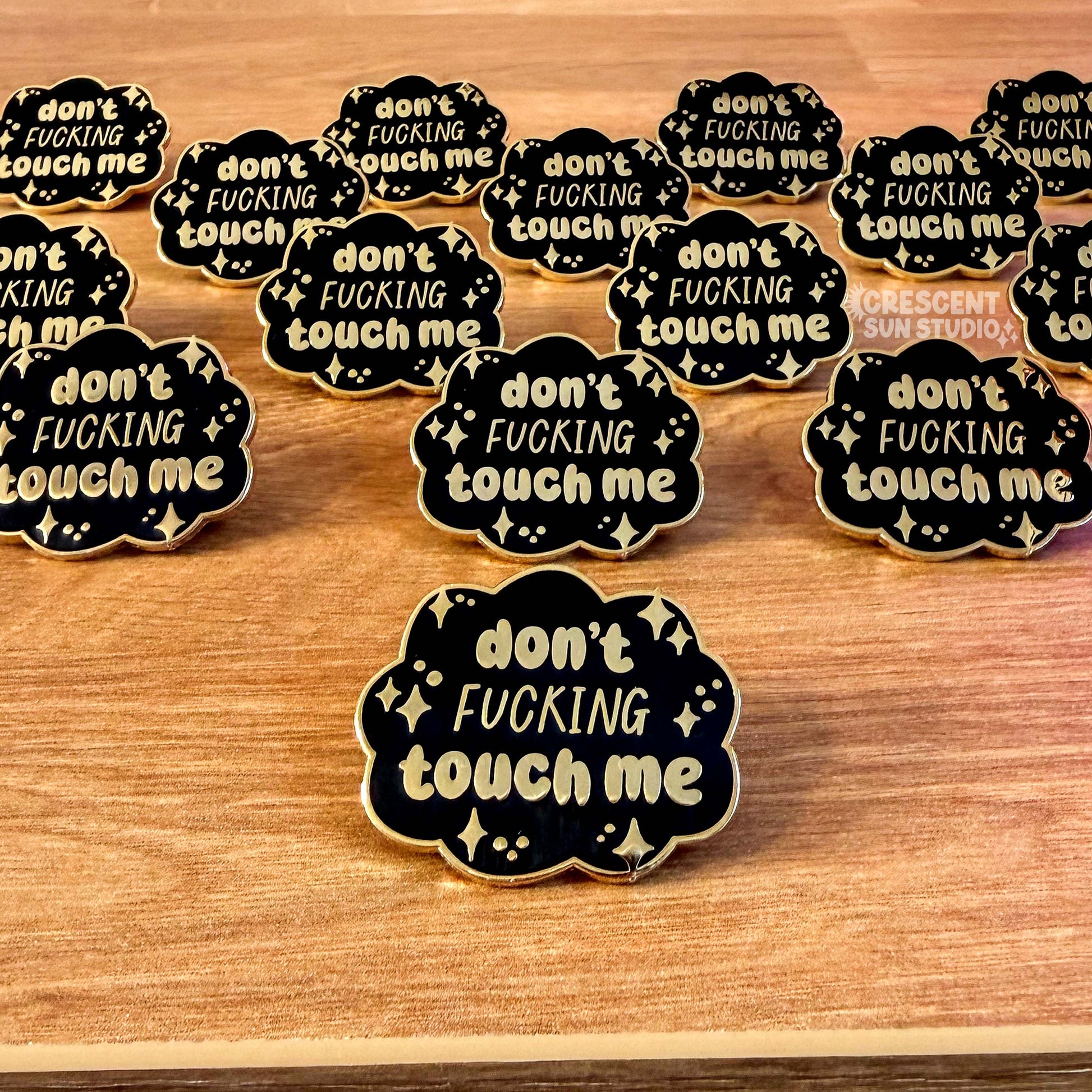 A group of cloud-shaped black pins with gold lettering that reads "don't fucking touch me" surrounded by little stars.