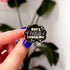 A hand holds a small cloud-shaped black pin with gold lettering that reads "don't fucking touch me" surrounded by little stars.