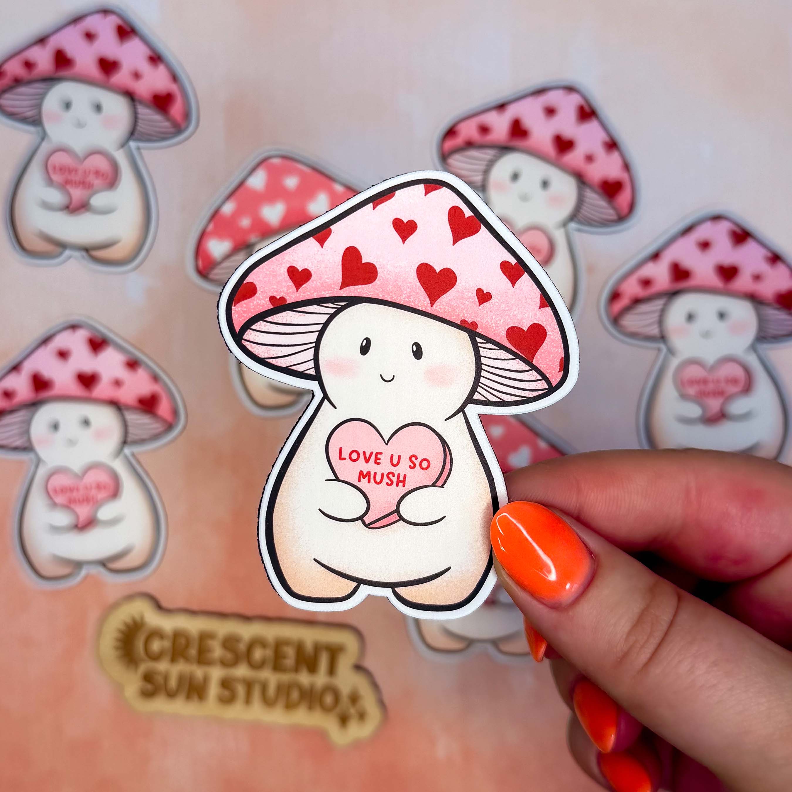 In this image, a hand holds one Dovey magnet close to the camera and there are other Dovey magnets in the background.

Dovey is a happy and plump cream mushroom character wearing a pink cap with red hearts. They are holding a pink candy heart that reads "Love U So Mush" in red lettering.