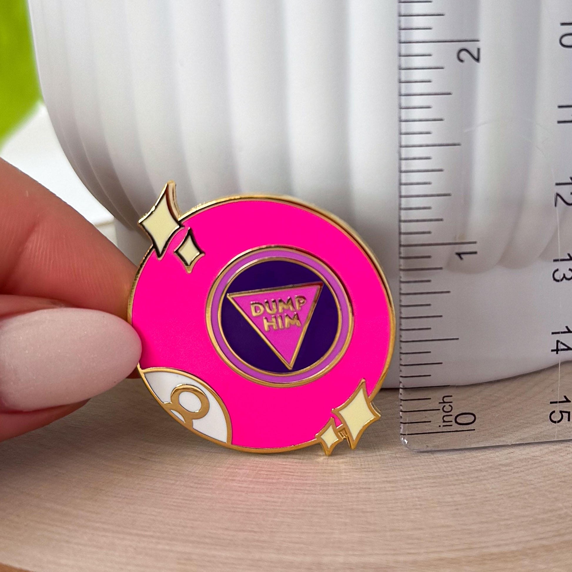 This bright pink fortune-telling ball inspired pin says "dump him" in the center of the ball. There are also two sets of stars on either side of the pin and part of the letter 8 is visible at the bottom left of the pin, as if curving around the ball.

In this image, the pin is placed next to a clear ruler, showing its height of roughly 1.25 inches.
