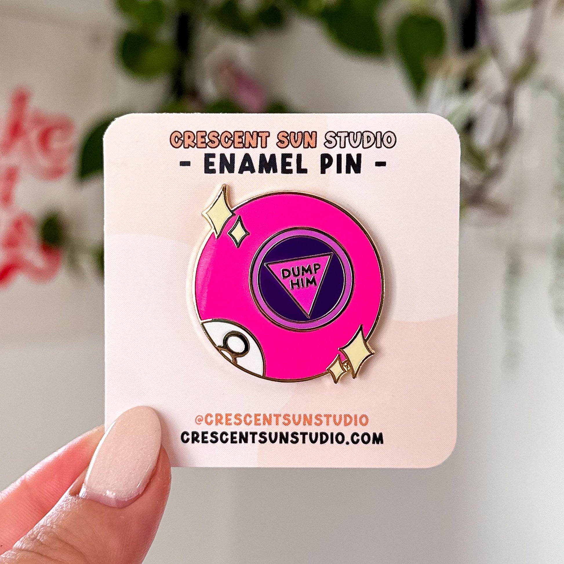 This bright pink 8 ball inspired pin says "dump him" in the center of the ball. There are also two sets of stars on either side of the pin and part of the letter 8 is visible at the bottom left of the pin, as if curving around the ball.

A hand holds the pin, displayed on a light colored card, close to the camera.