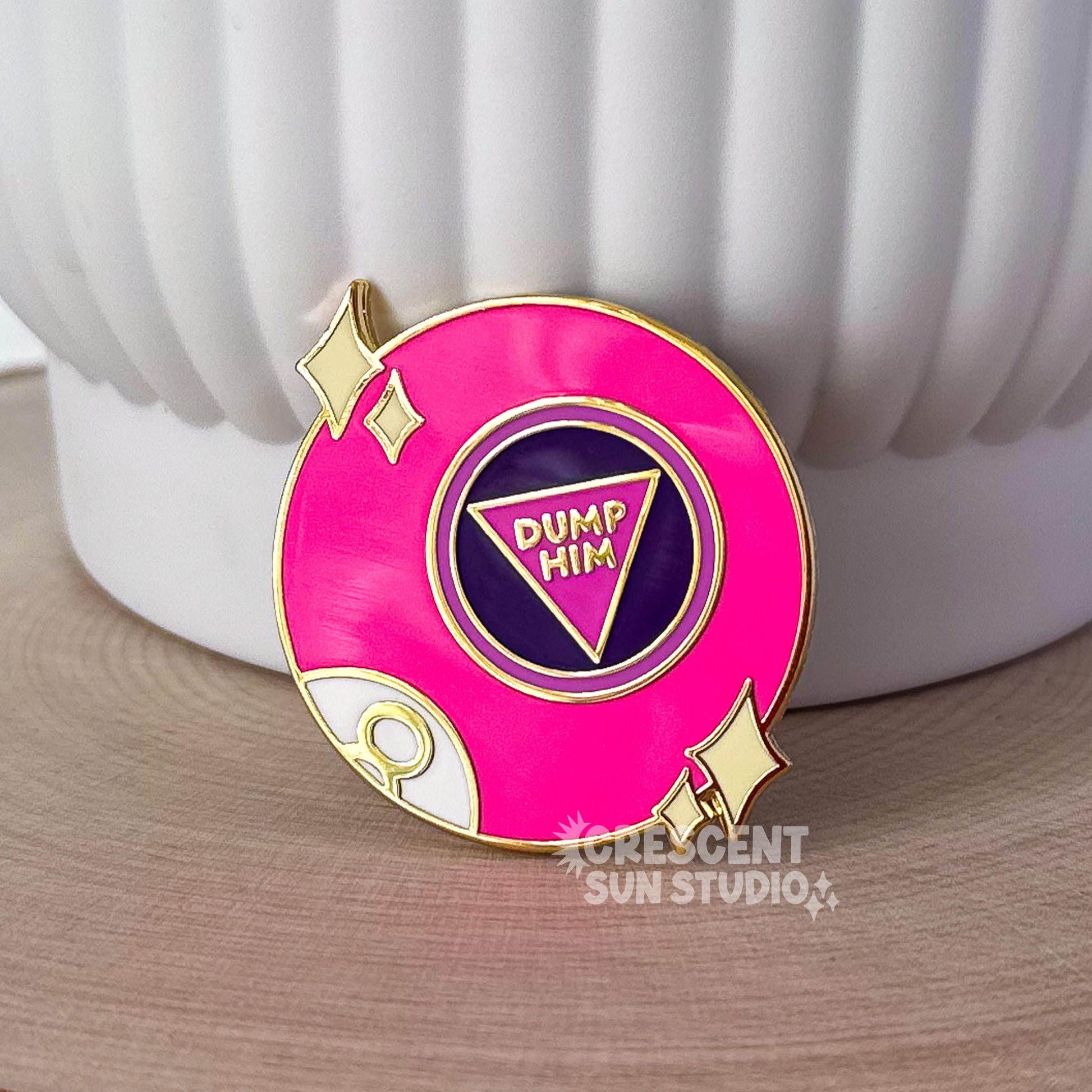This bright pink fortune telling ball enamel pin says "dump him" in the center of the ball. There are also two sets of stars on either side of the pin and part of the letter 8 is visible at the bottom left of the pin, as if curving around the ball.

In this image, the pin leans against a large white plant pot.