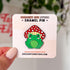An enamel pin in the shape of a happy green frog wearing a red and white mushroom for a hat. 

In this image, the pin is displayed on a light colored card.