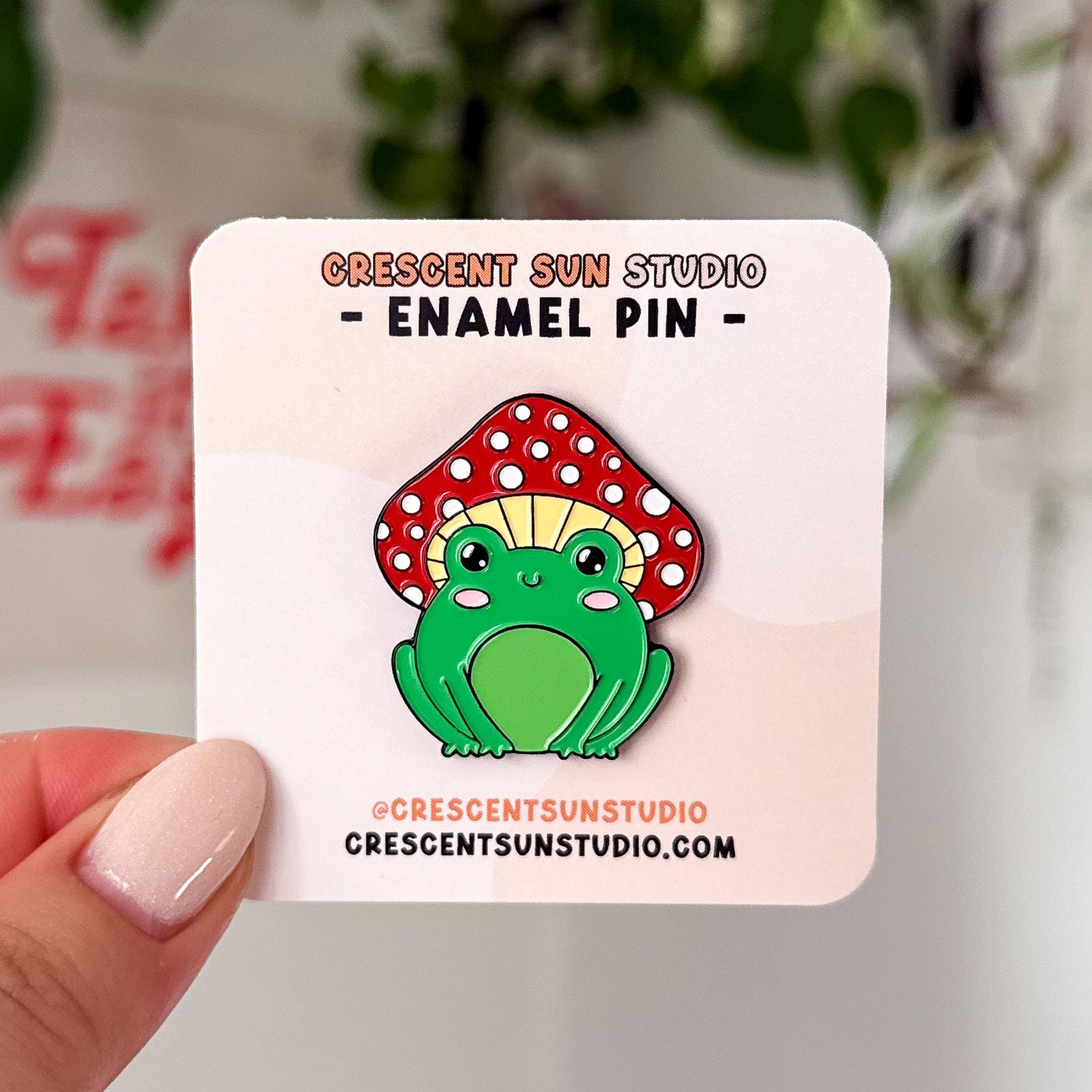 An enamel pin in the shape of a happy green frog wearing a red and white mushroom for a hat. 

In this image, the pin is displayed on a light colored card.