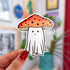 A sticker in the shape of a ghost wearing an orange mushroom top for a hat. The mushroom has dark dots on it and the ghostie has a smiling face.