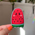 A sticker that looks like a red seeded watermelon slice shaped like a ghost is held close to the camera.