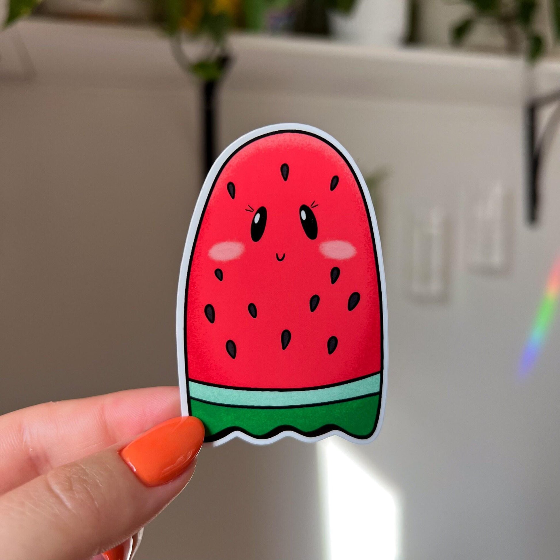A sticker that looks like a red seeded watermelon slice shaped like a ghost is held close to the camera.