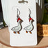 A closeup of pair of dangle earrings, each featuring a side view of a white goose mid-stride. Each goose wears a red and white spotted mushroom cap on its head. The earrings are displayed on a white card.