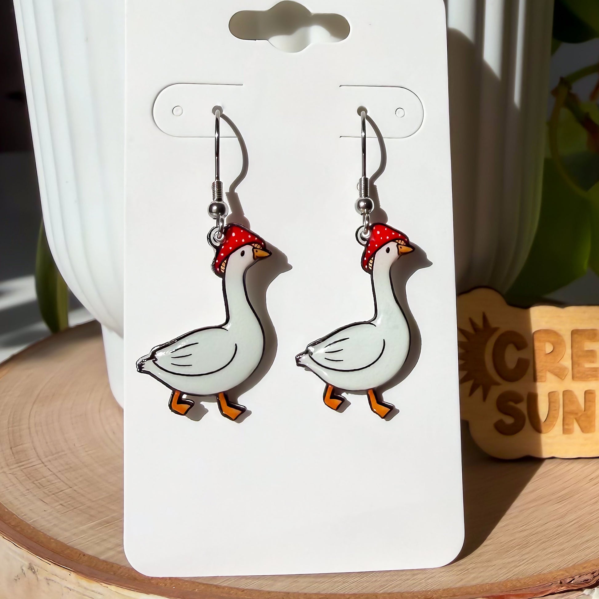 A closeup of pair of dangle earrings, each featuring a side view of a white goose mid-stride. Each goose wears a red and white spotted mushroom cap on its head. The earrings are displayed on a white card.