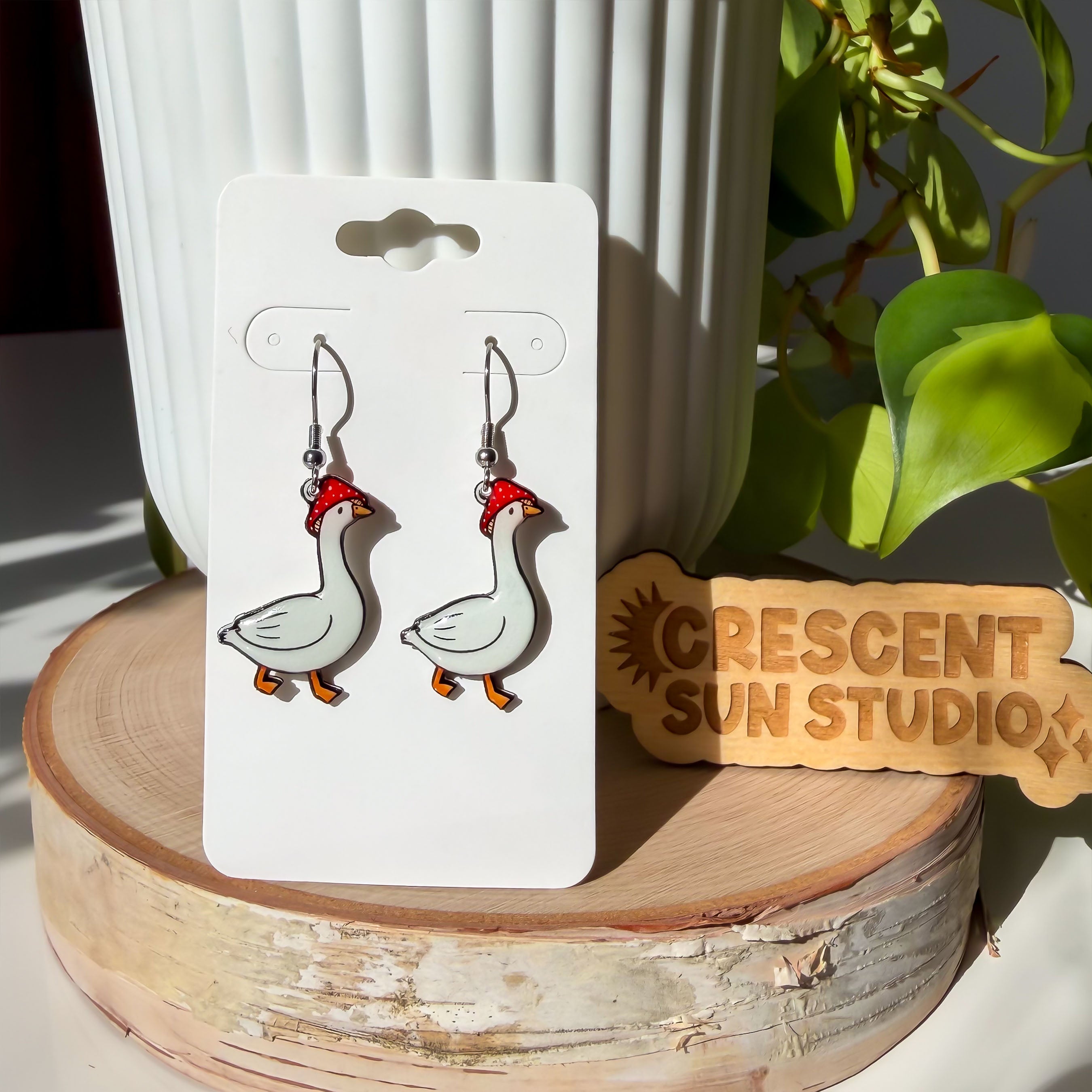 A pair of dangle earrings, each featuring a side view of a white goose mid-stride. Each goose wears a red and white spotted mushroom cap on its head. 

The earrings are displayed on a white card.