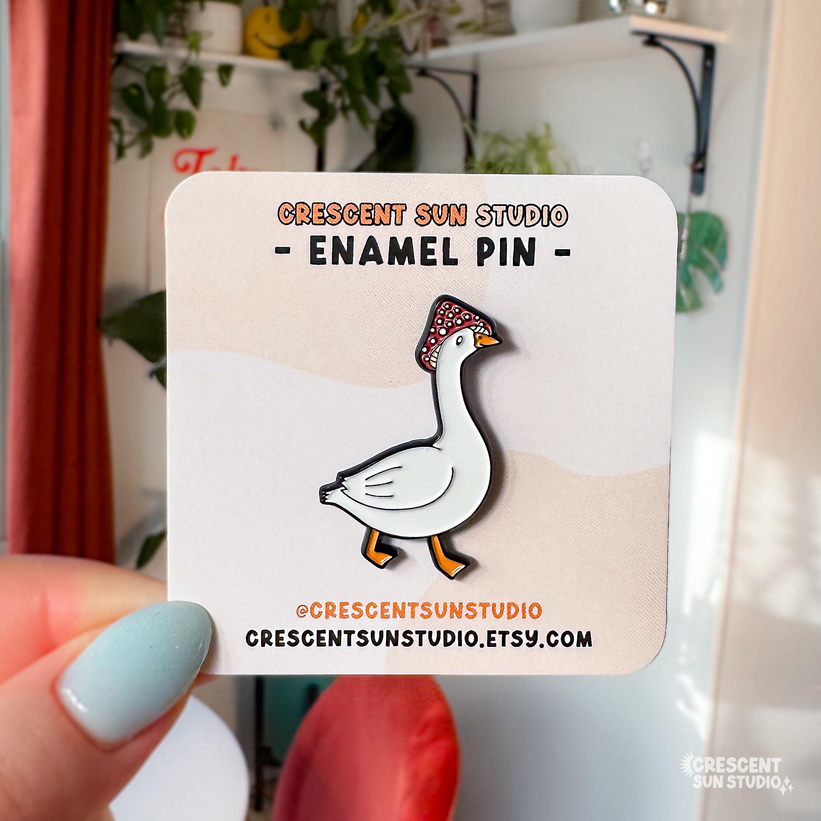 Image shows an enamel pin that is a side profile of a white goose wearing a red and white spotted mushroom for a hat. The pin is displayed on a card that says "Crescent Sun Studio" and "enamel pin"