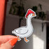 A closeup of our goose mushroom sticker features a very silly, but unbothered, white goose wearing a red and white spotted mushroom cap on its head. The sticker shows the white goose's side profile and the bird is also mid-step.
