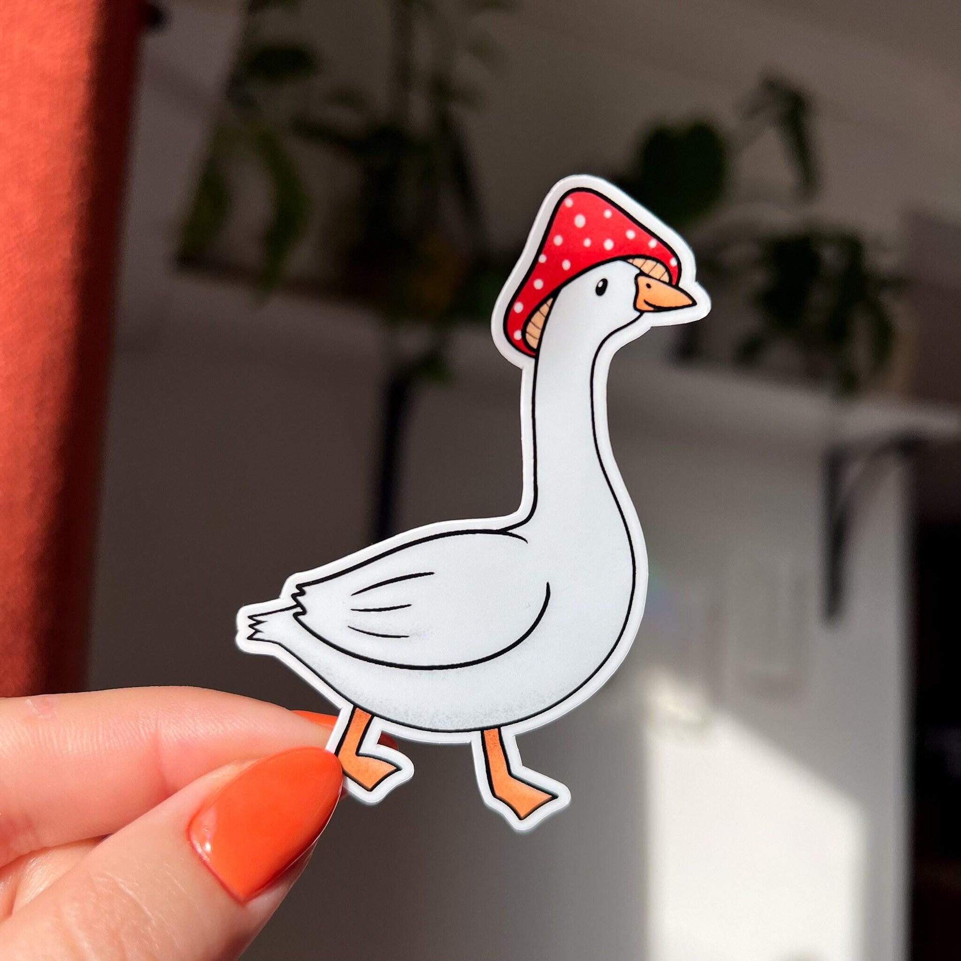 A closeup of our goose mushroom sticker features a very silly, but unbothered, white goose wearing a red and white spotted mushroom cap on its head. The sticker shows the white goose's side profile and the bird is also mid-step.