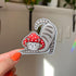 A sticker in the shape of a cat wearing a mushroom hat is held close to the camera. The cat is a grey tabby (medium grey with darker grey stripes) and has a sweet expression on his face. His red mushroom hat with white spots has spots for his ears to poke through. The cat is in a stretching pose with his tail curling up behind him. 

In this image, a woman (off-camera) holds the sticker in her hand. Her thumb nail is painted a bright coral orange.