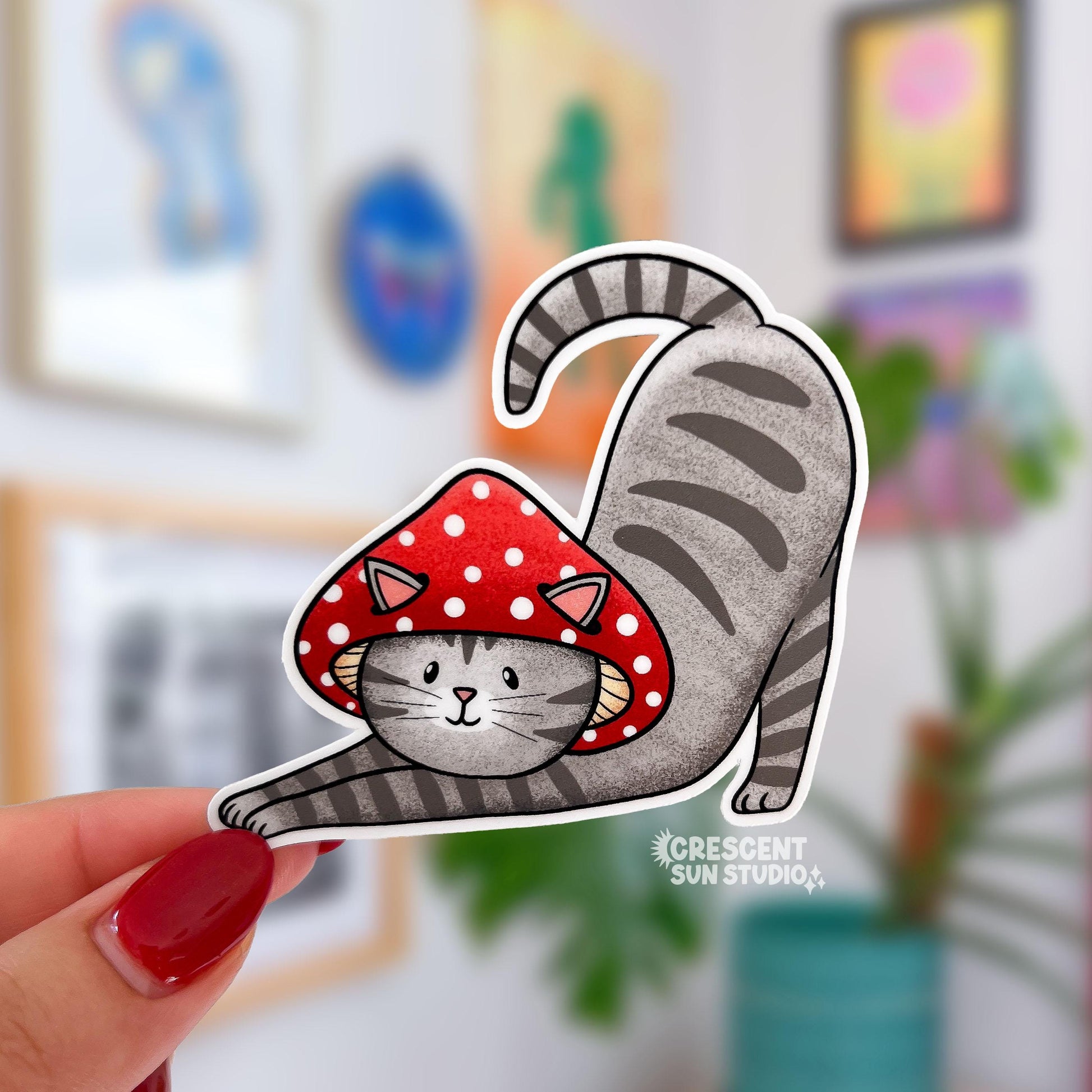A sticker in the shape of a cat wearing a mushroom hat is held close to the camera. The cat is a grey tabby (medium grey with darker grey stripes) and has a sweet expression on his face. His red mushroom hat with white spots has spots for his ears to poke through. The cat is in a stretching pose with his tail curling up behind him.

In this image, a person holds the cat-shaped sticker up close to the camera.