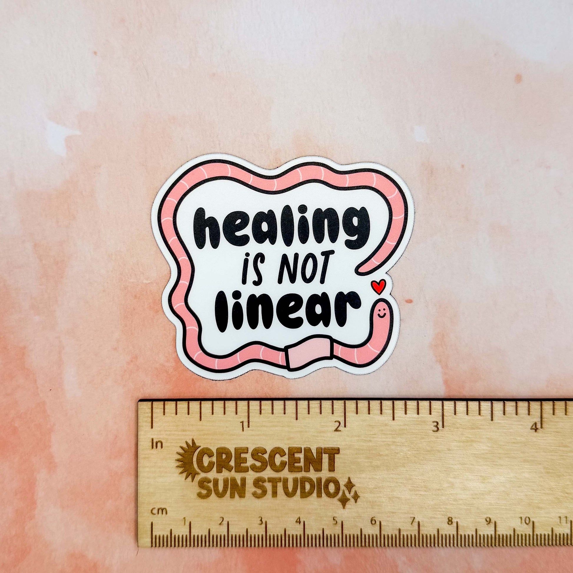 The magnet features bold black text surrounded by a friendly segmented worm. The worm's head and his tail do not connect, instead, there is a tiny heart separating the two. In this image, the magnet sits above a wooden ruler, showing its width of 2.4 inches.