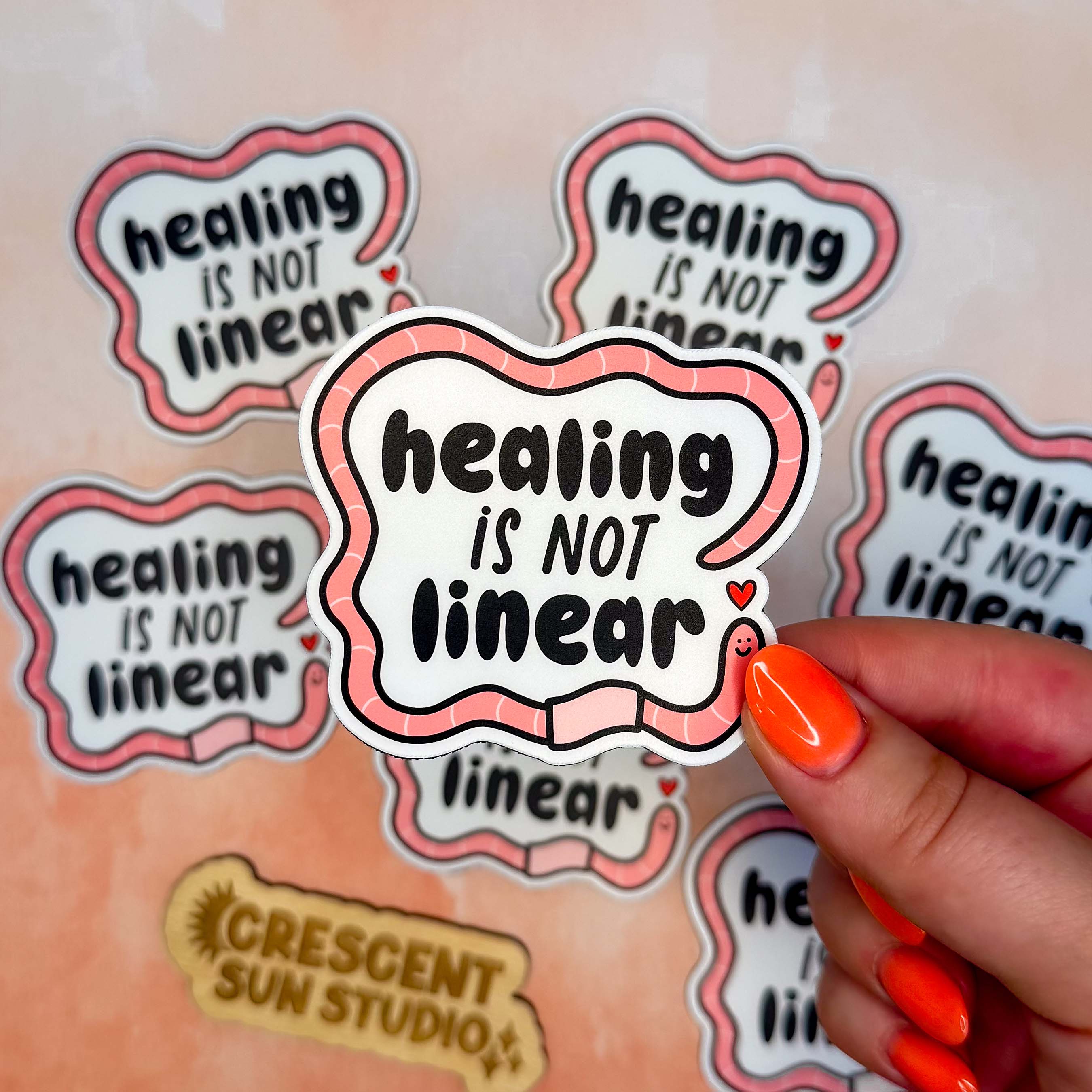 The design features bold black text surrounded by a friendly segmented worm. The worm's head and his tail do not connect, instead, there is a tiny heart separating the two. In this image, one magnet is held close to the camera by a woman's hand. There are other scattered magnets on the table below the central image.