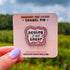 The design features bold black text surrounded by a friendly segmented worm. The text reads, "healing is not linear." The worm's head and his tail do not connect, instead, there is a tiny heart separating the two. In this image, the pin is on a display card and held close to the camera.