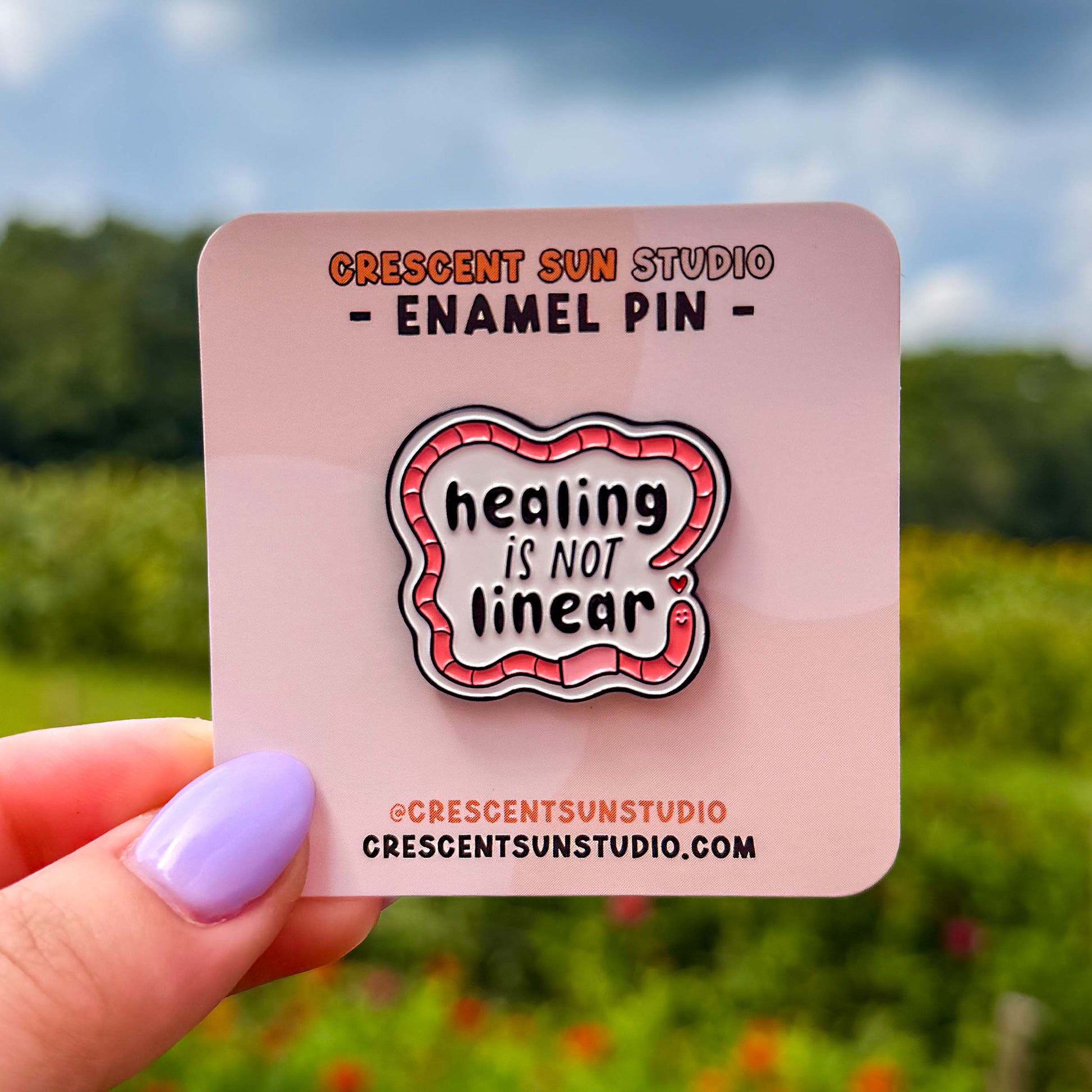 The design features bold black text surrounded by a friendly segmented worm. The text reads, "healing is not linear." The worm's head and his tail do not connect, instead, there is a tiny heart separating the two. In this image, the pin is on a display card and held close to the camera.