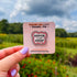 The design features bold black text surrounded by a friendly segmented worm. The text reads, "healing is not linear." The worm's head and his tail do not connect, instead, there is a tiny heart separating the two. In this image, the pin is on a display card and held close to the camera.