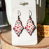 A pair of earrings dangle from silver-colored hooks. The earrings resemble ghosts with eyes and are covered in little red hearts.