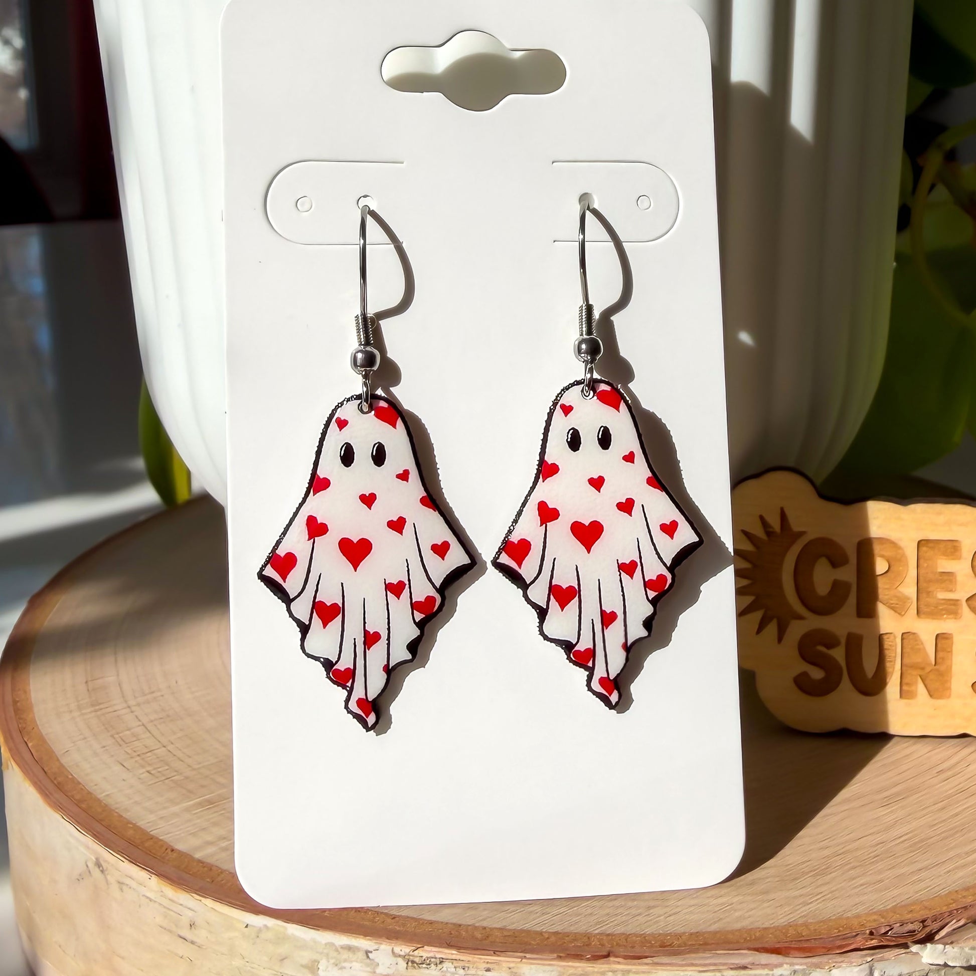 A pair of earrings dangle from silver-colored hooks. The earrings resemble ghosts with eyes and are covered in little red hearts.