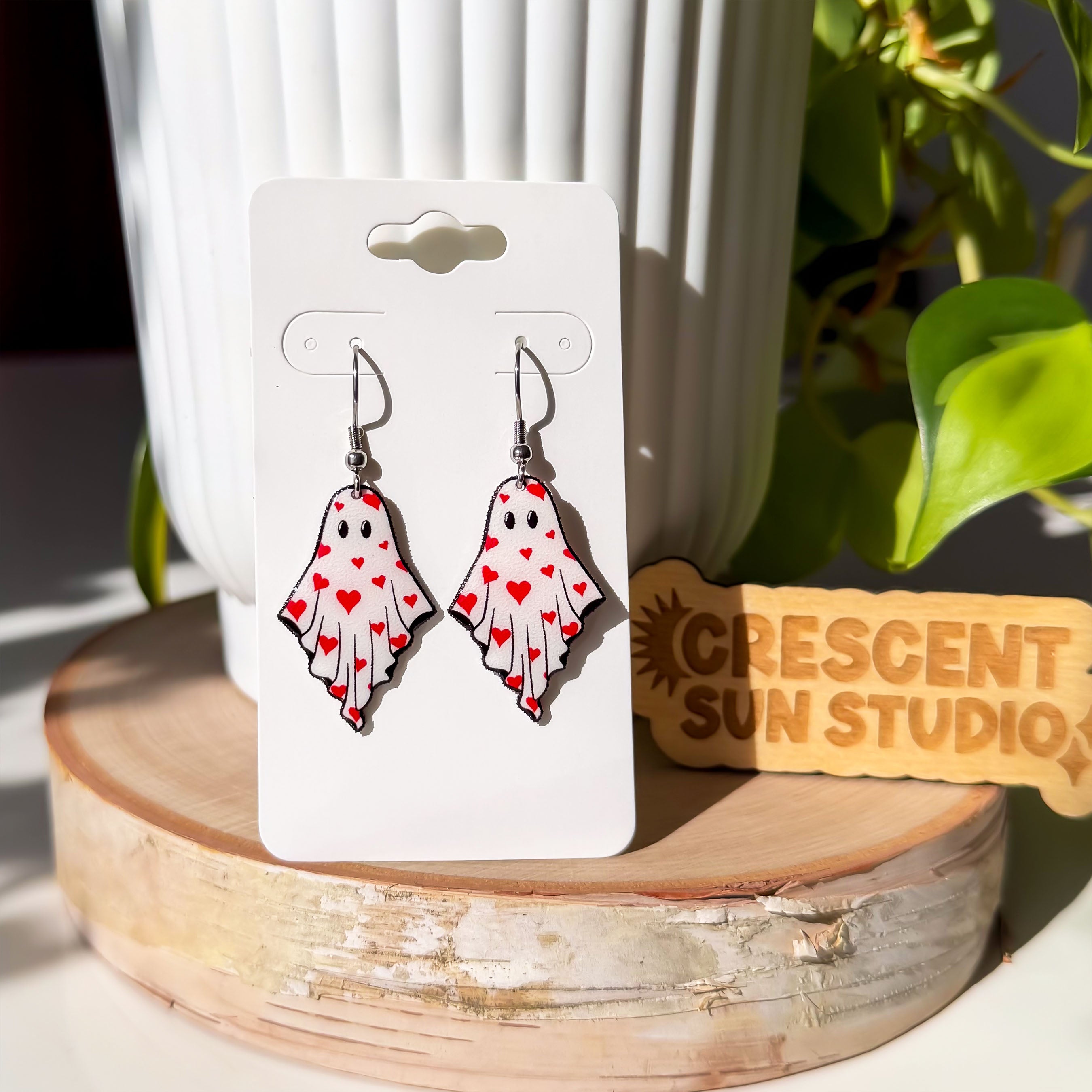 A pair of earrings dangle from silver-colored hooks. The earrings resemble ghosts with eyes and are covered in little red hearts.