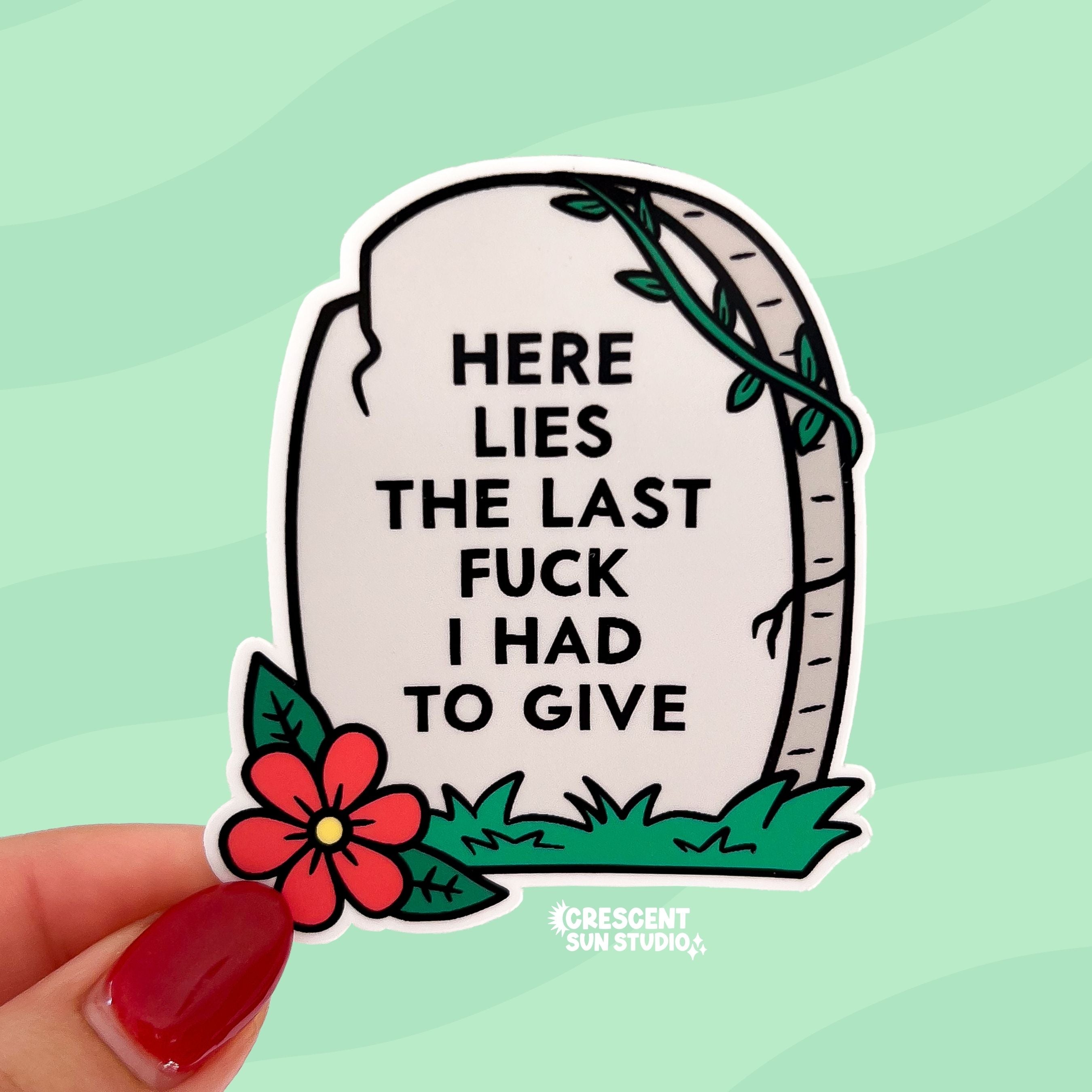 The sticker is in the shape of a light colored tombstone with slight cracks at various points. There is a red flower at the bottom left corner, green green grass, and then a green vine at the top right corner. Written on the gravestone is "here lies the last fuck I had to give" in all caps.