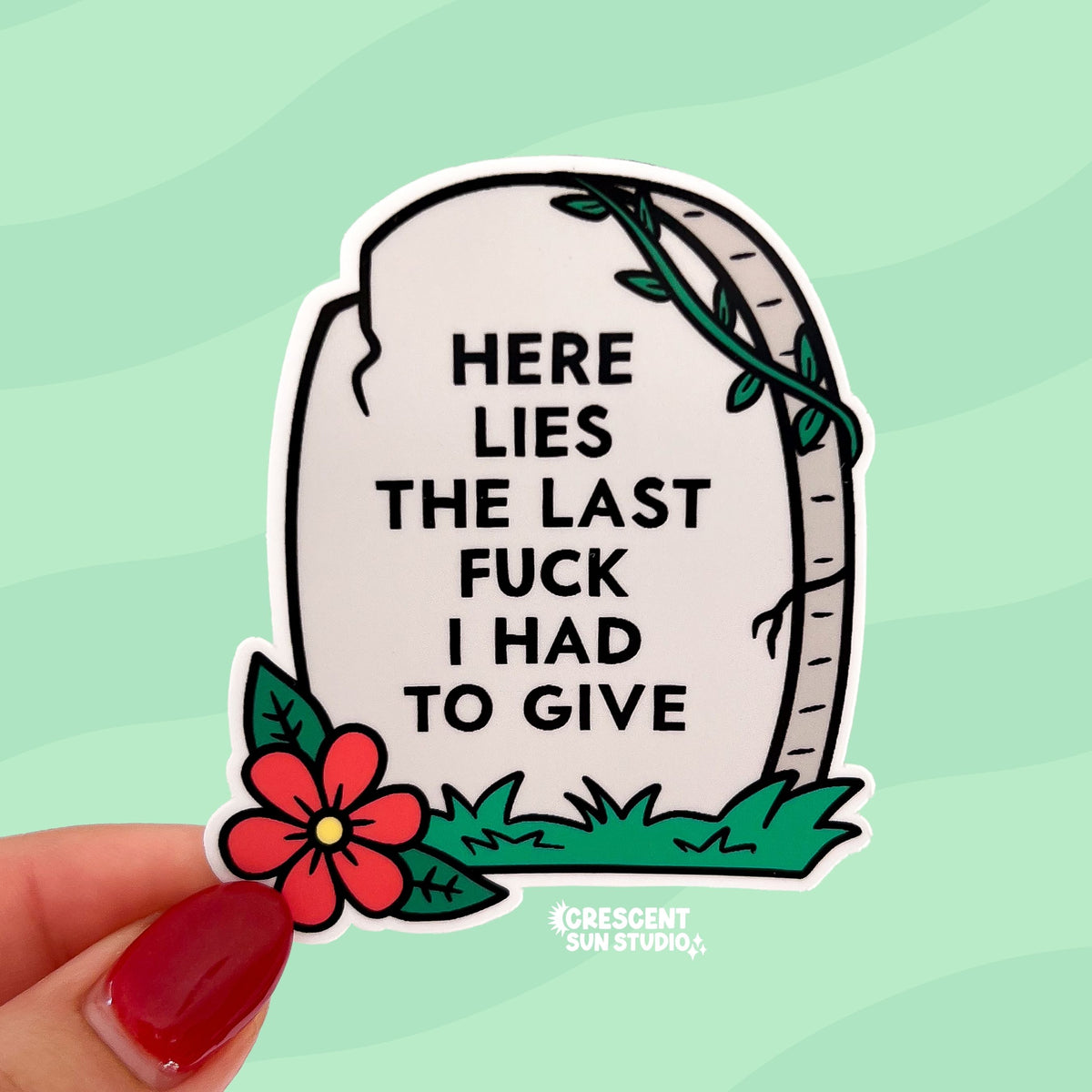 The sticker is in the shape of a light colored tombstone with slight cracks at various points. There is a red flower at the bottom left corner, green green grass, and then a green vine at the top right corner. Written on the gravestone is "here lies the last fuck I had to give" in all caps.