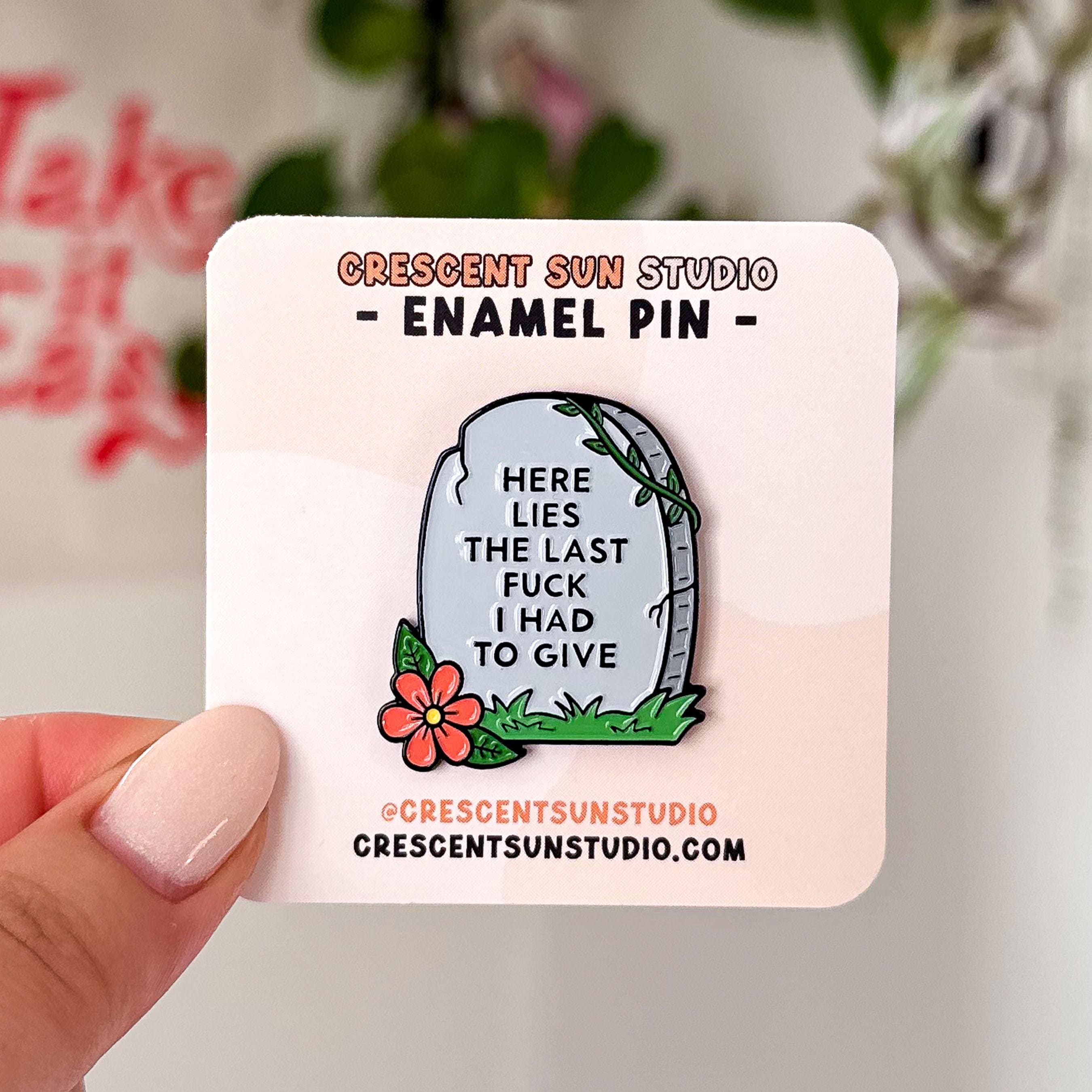 The pin is in the shape of a light colored tombstone with slight cracks at various points. There is a red flower at the bottom left corner, green green grass, and then a green vine at the top right corner. Written on the gravestone is "here lies the last fuck I had to give" in all caps.

In this image, the pin is on a light-colored display card and is held up close to the camera.