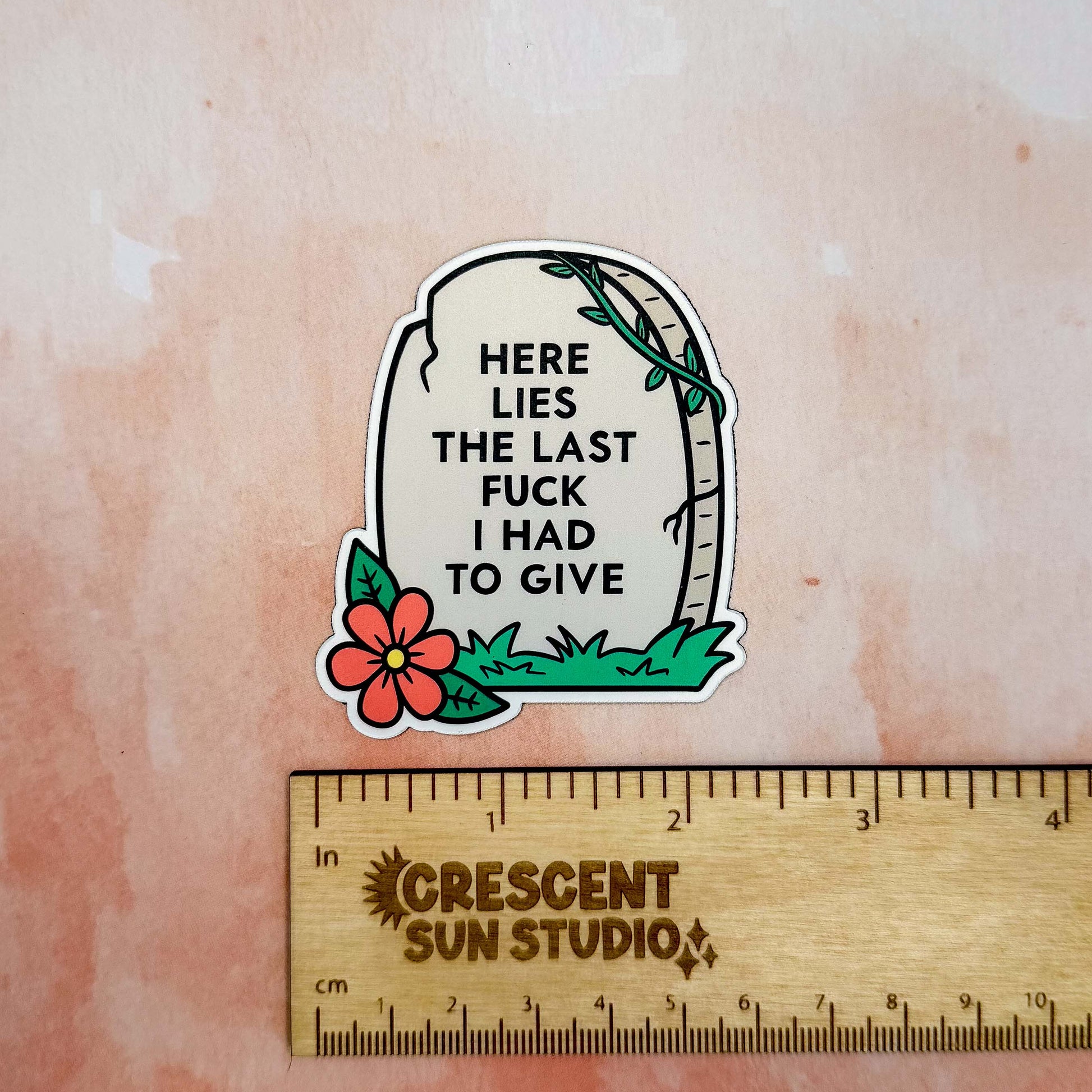 The magnet is in the shape of a light colored tombstone with slight cracks at various points. There is a red flower at the bottom left corner, green green grass, and then a green vine at the top right corner. Written on the tombstone is "here lies the last fuck I had to give" in all caps.

In this image, the magnet is above a wooden ruler that shows its width of 2.3 inches.
