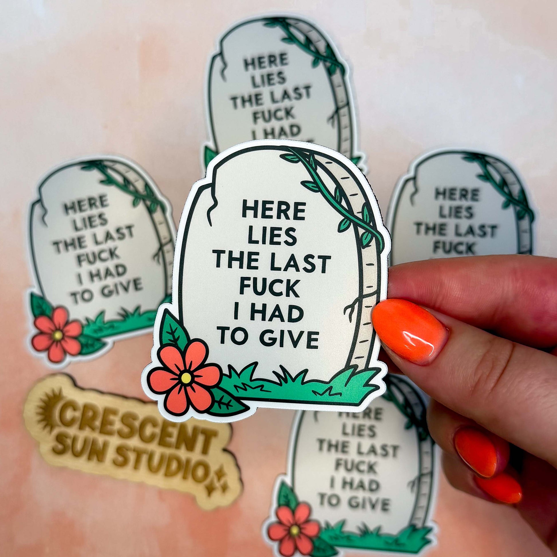 The magnet is in the shape of a light colored tombstone with slight cracks at various points. There is a red flower at the bottom left corner, green green grass, and then a green vine at the top right corner. Written on the tombstone is "here lies the last fuck I had to give" in all caps.

In this image, a hand holds one magnet close to the camera and there are other magnets in the background.