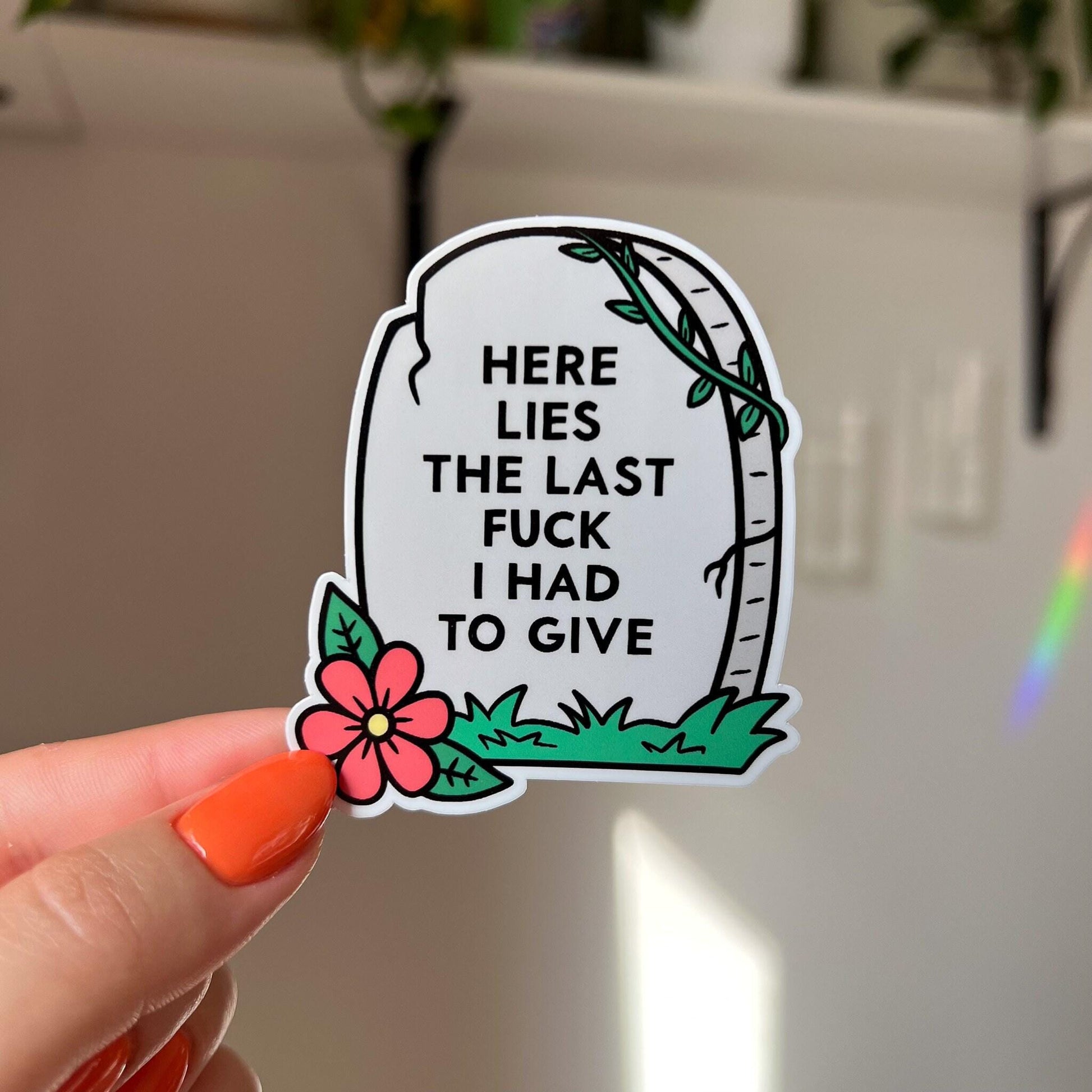 The sticker is in the shape of a light colored tombstone with slight cracks at various points. There is a red flower at the bottom left corner, green green grass, and then a green vine at the top right corner. Written on the gravestone is "here lies the last fuck I had to give" in all caps.

In this image, a hand holds the sticker close to the camera against a light and bright white wall in the background.