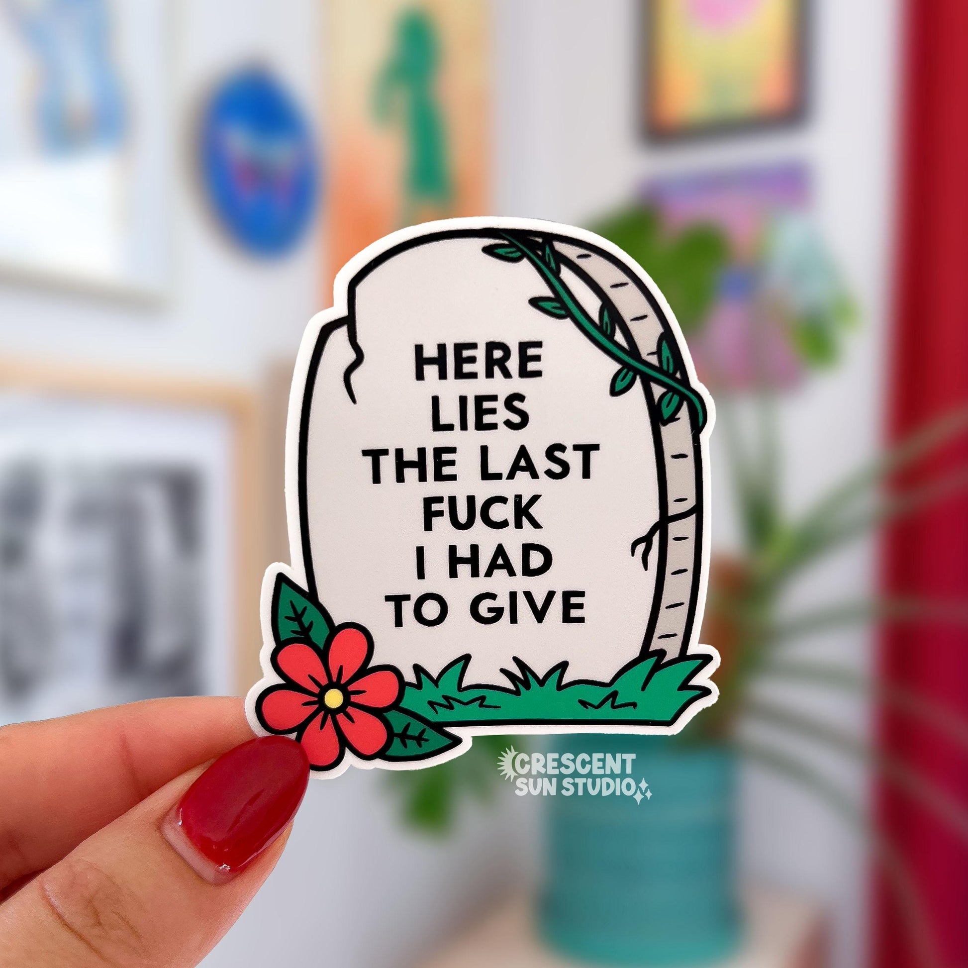 The sticker is in the shape of a light colored tombstone with slight cracks at various points. There is a red flower at the bottom left corner, green green grass, and then a green vine at the top right corner. Written on the gravestone is "here lies the last fuck I had to give" in all caps.

In this image, a hand holds one sticker close to the camera and there are plants in the background.