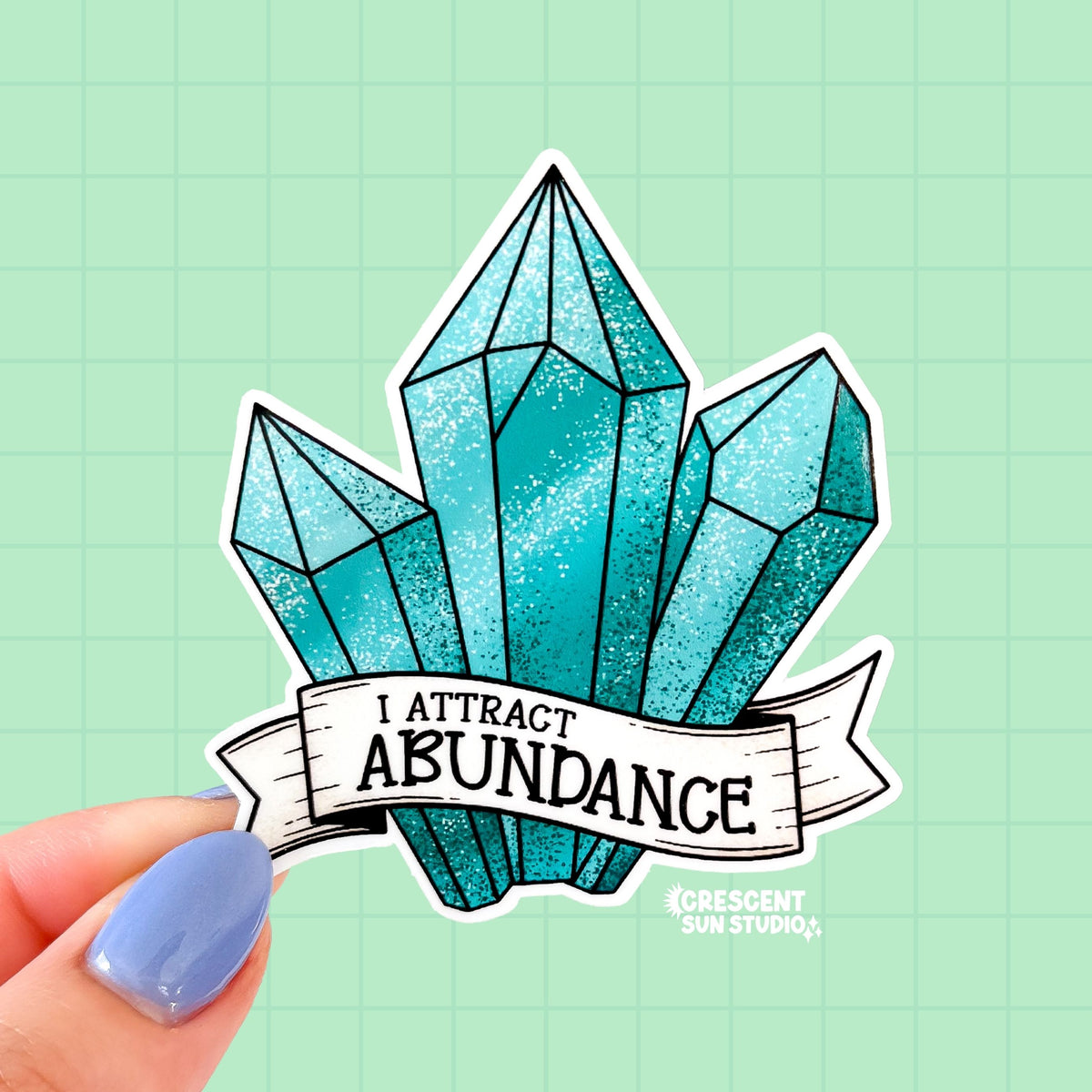 A sticker in the shape of three turquoise shaded crystals. At the bottom is a white banner with black text that reads "I attract abundance"