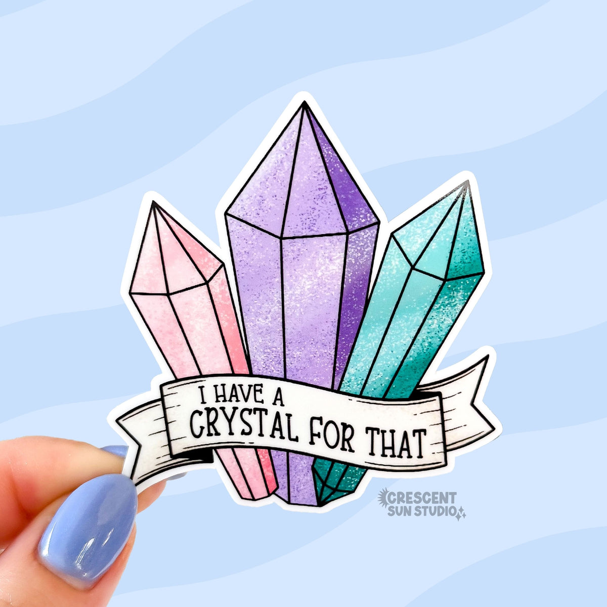 A sticker in the shape of three pastel shaded crystals in pink, purple, and turquoise. At the bottom is a white banner with black text that reads "I have a crystal for that"