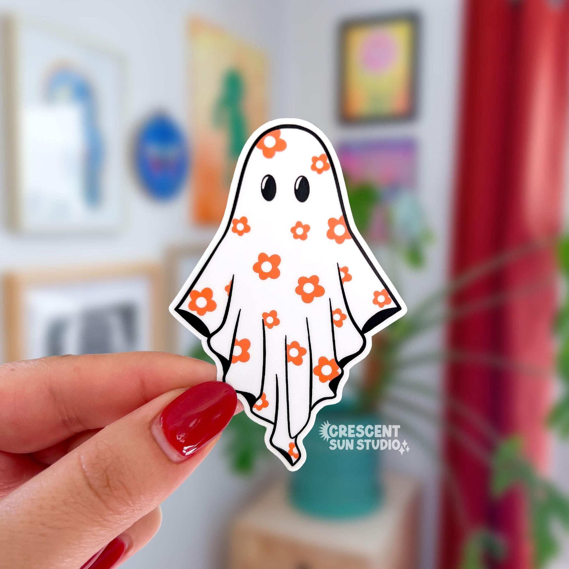 Hand holding a ghost-shaped sticker with floral patterns against a blurred indoor background