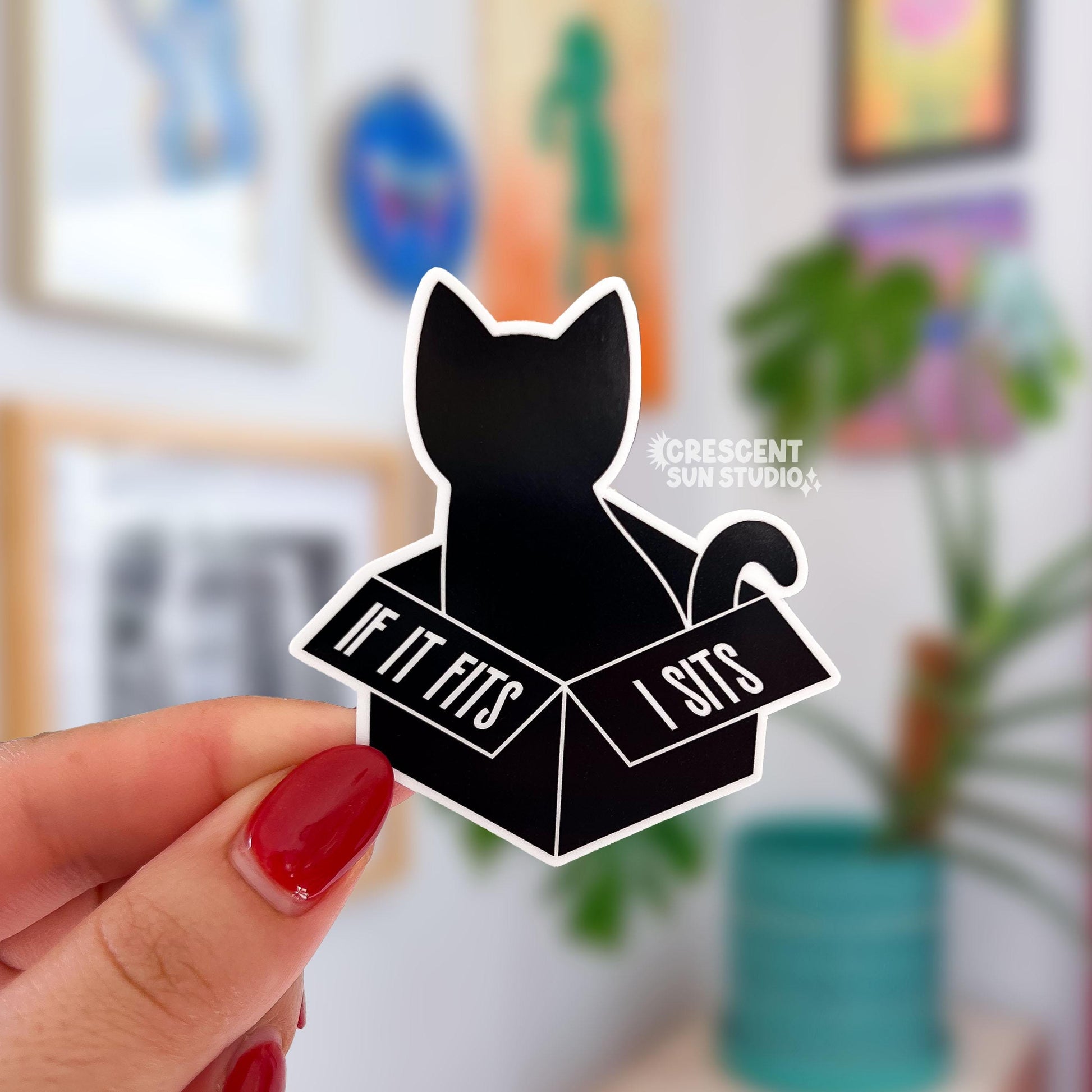 A sticker in the shape of a cat silhouette sitting inside of a box. Both the cat and the box are black and outlined with white. On the open flaps of the box it reads "if it fits" on the left flap and on the right "I sits."